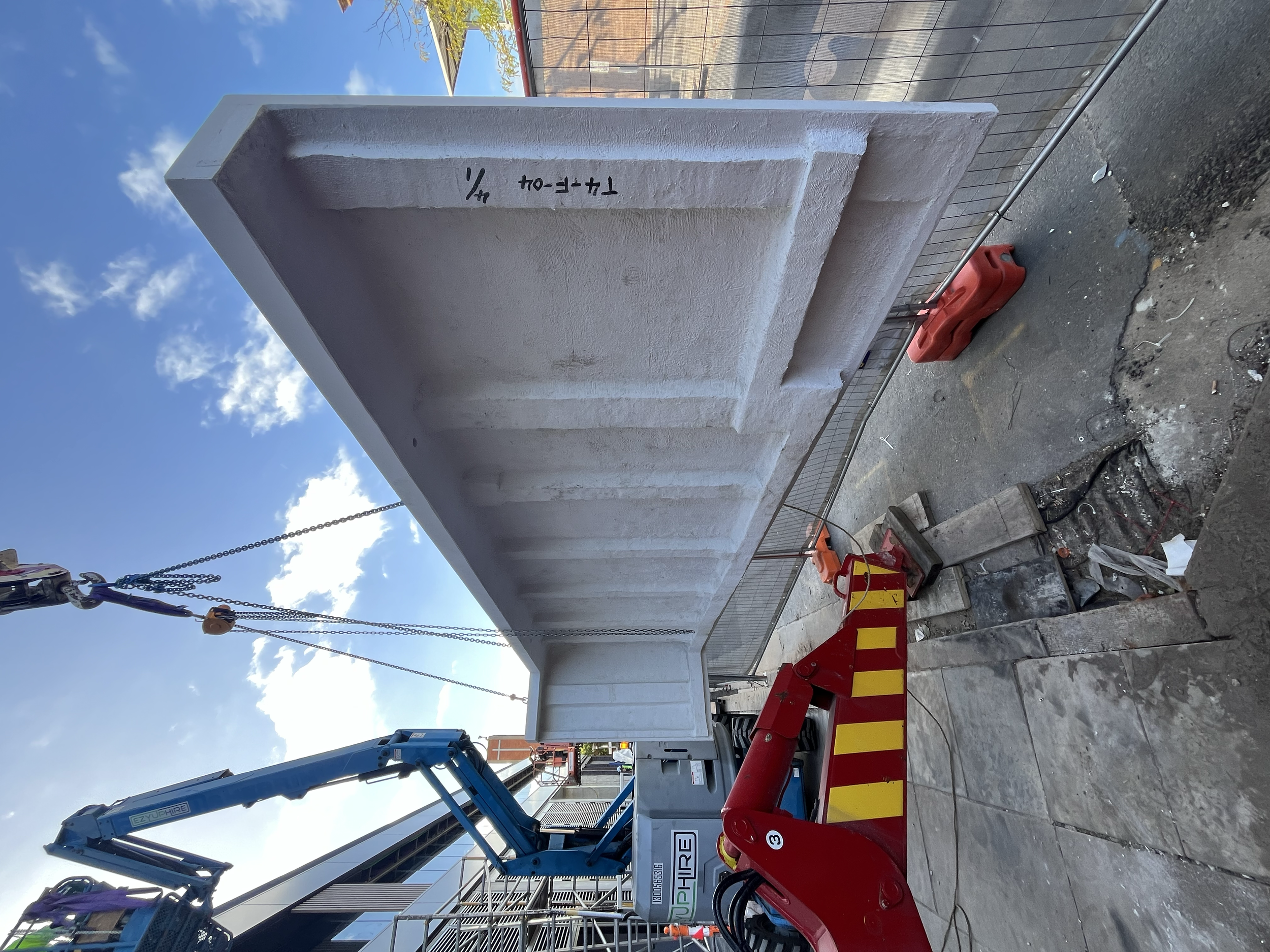 Construction site with a large white concrete structure, secured by orange barriers, a red hydraulic lift, and a blue crane arm against a partly cloudy sky.