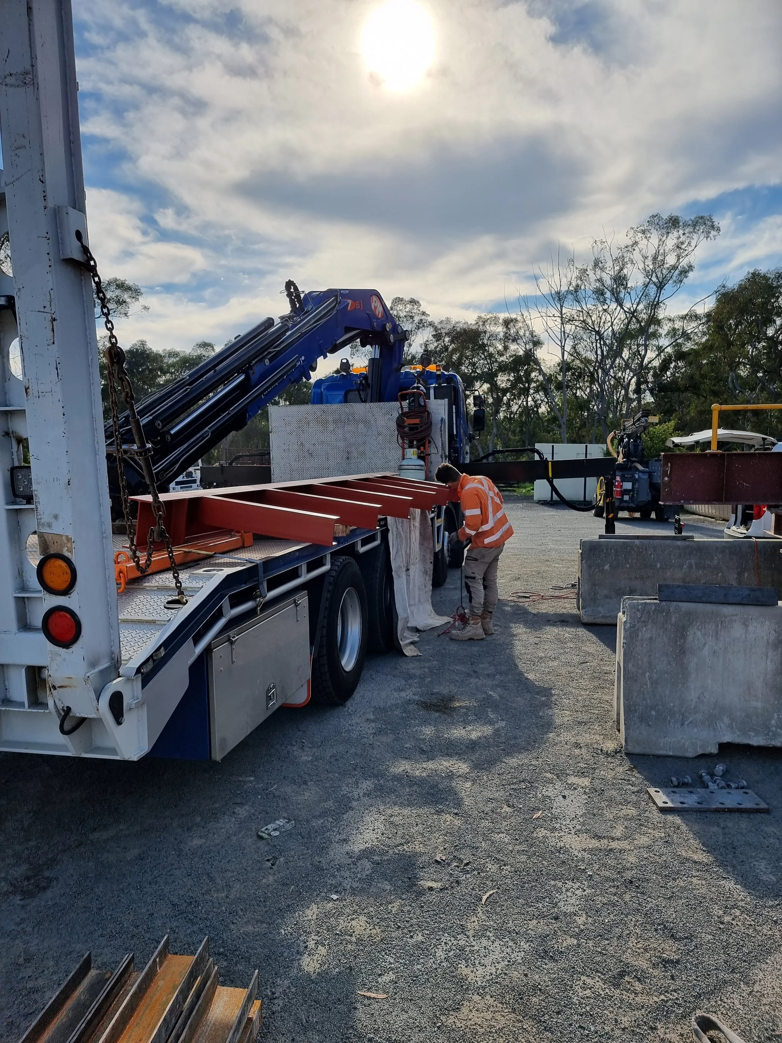 Worker inspecting or working on construction equipment at a construction site with a truck, metal beams, and barriers, under a partly cloudy sky during the day.