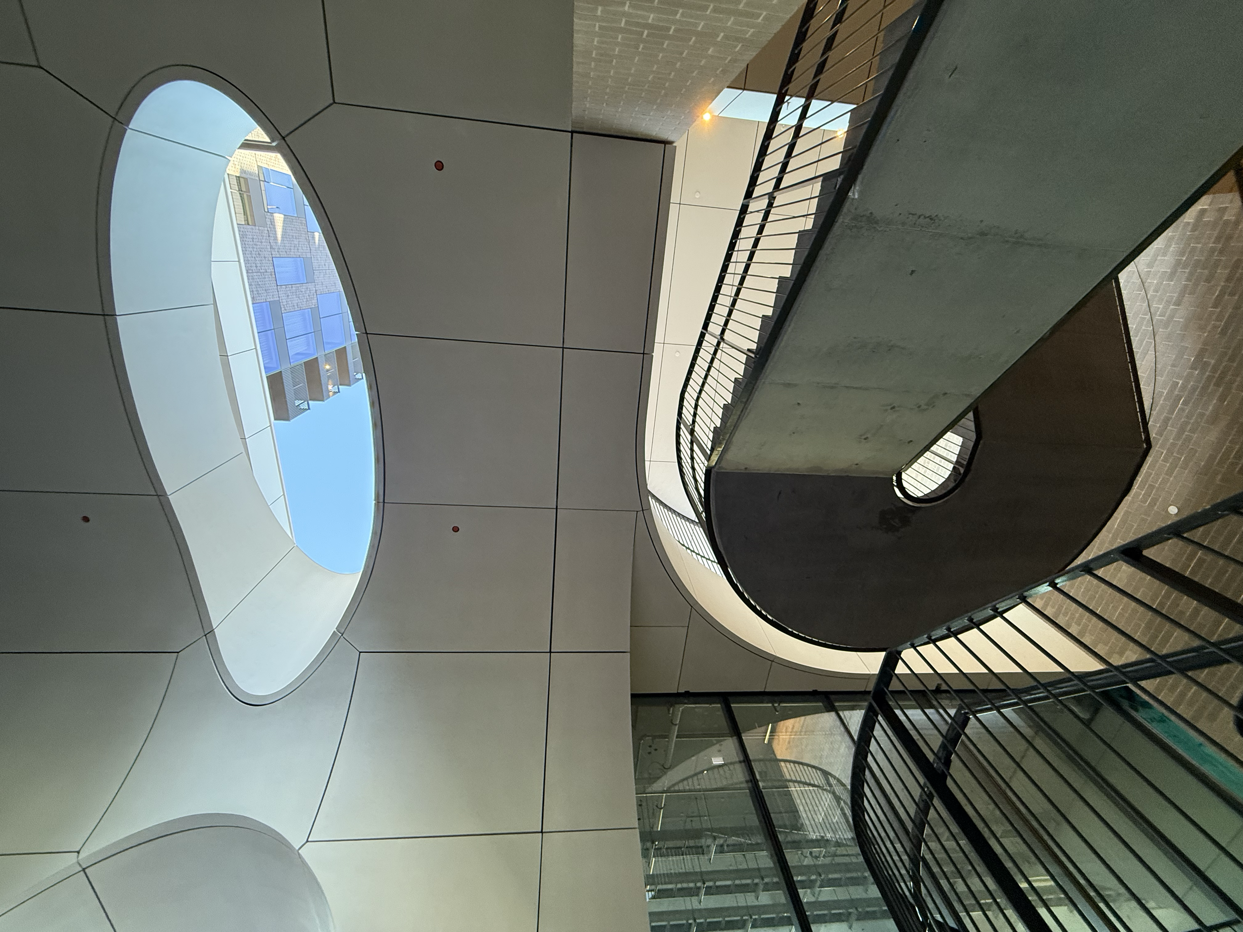 Interior view of a modern building with curved architectural elements, including a staircase with metal railings, and circular openings in the ceiling showing a blue sky and adjacent building.