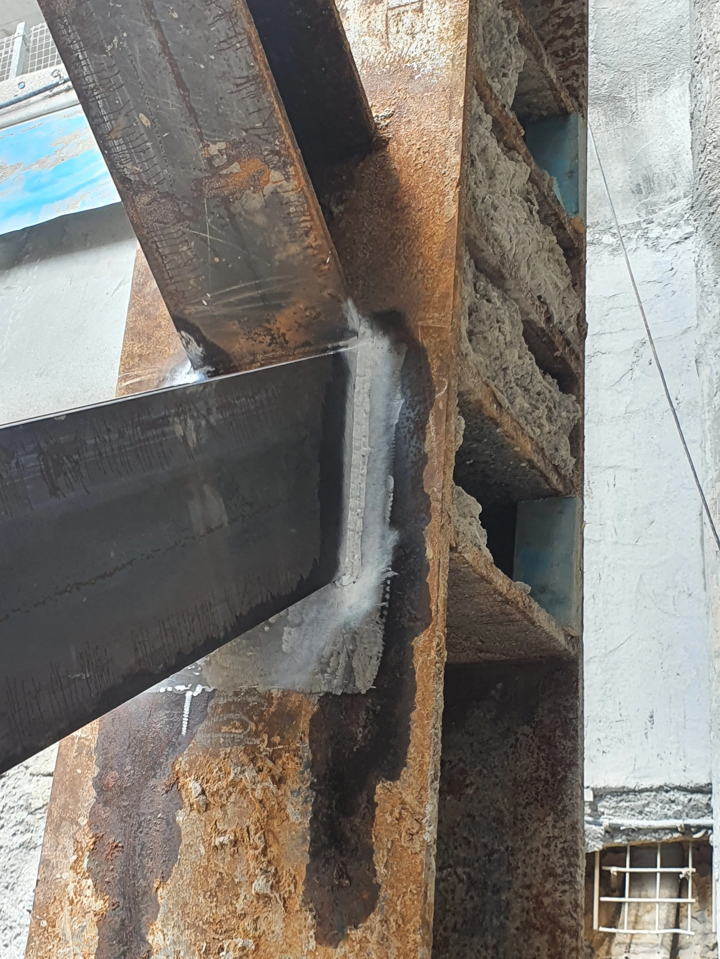 Close-up of a rusty metal structure with fresh welding, binding a steel beam to a larger metal frame, with a concrete wall in the background.