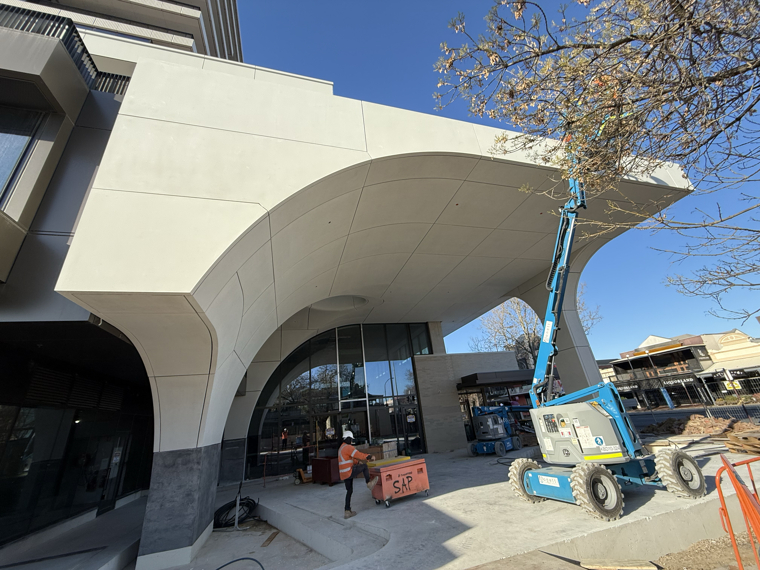 Construction workers in safety gear work outside a modern building with a unique arched entrance, using a blue lift to access the upper areas, surrounded by construction materials and fencing.