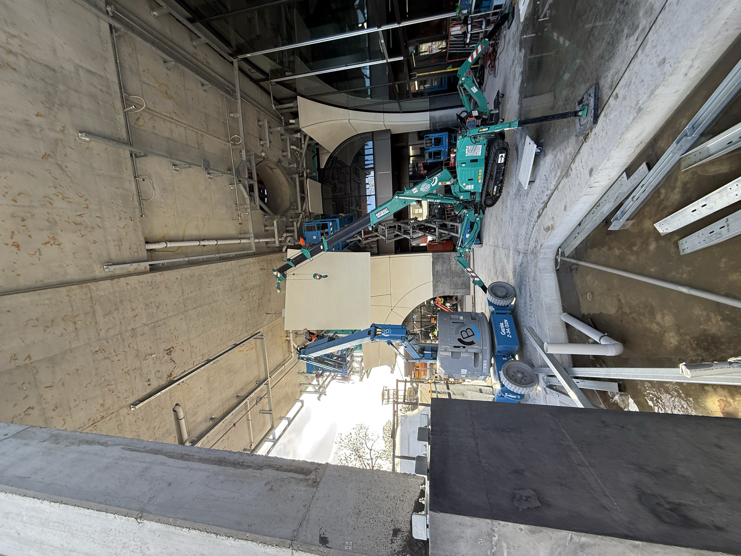 Construction site with a blue articulated boom lift extending over a partially built wall, with metal scaffolding, ladders, and construction materials around.