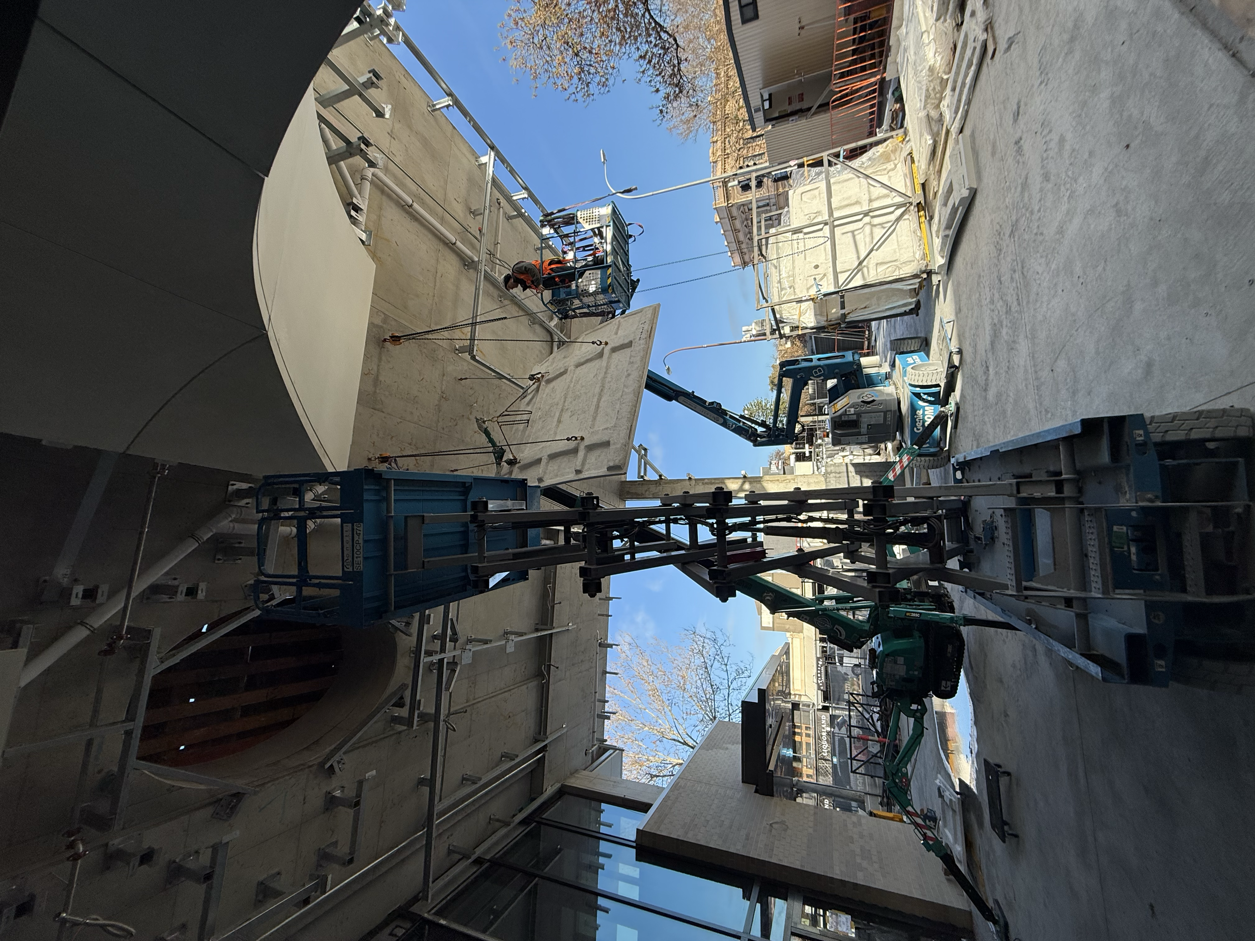 Construction site with heavy machinery and workers installing large exterior panels on a building, with blue sky in the background.