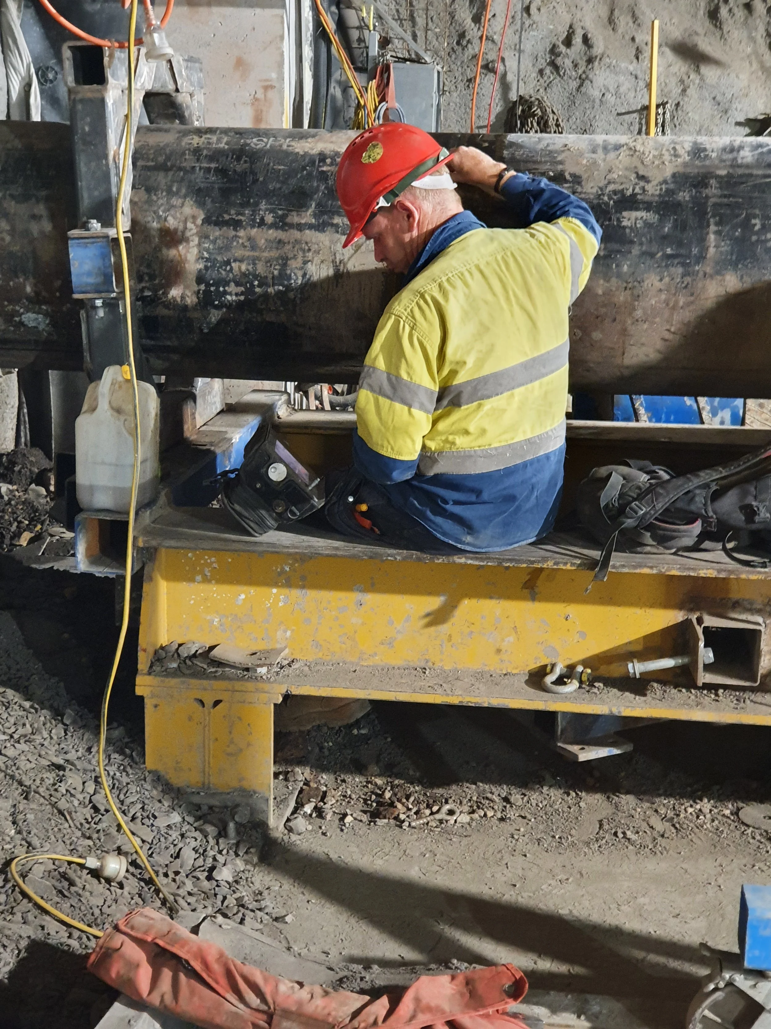 A construction worker wearing a red safety helmet and yellow high-visibility jacket working on a large pipeline or pipe on a construction site.