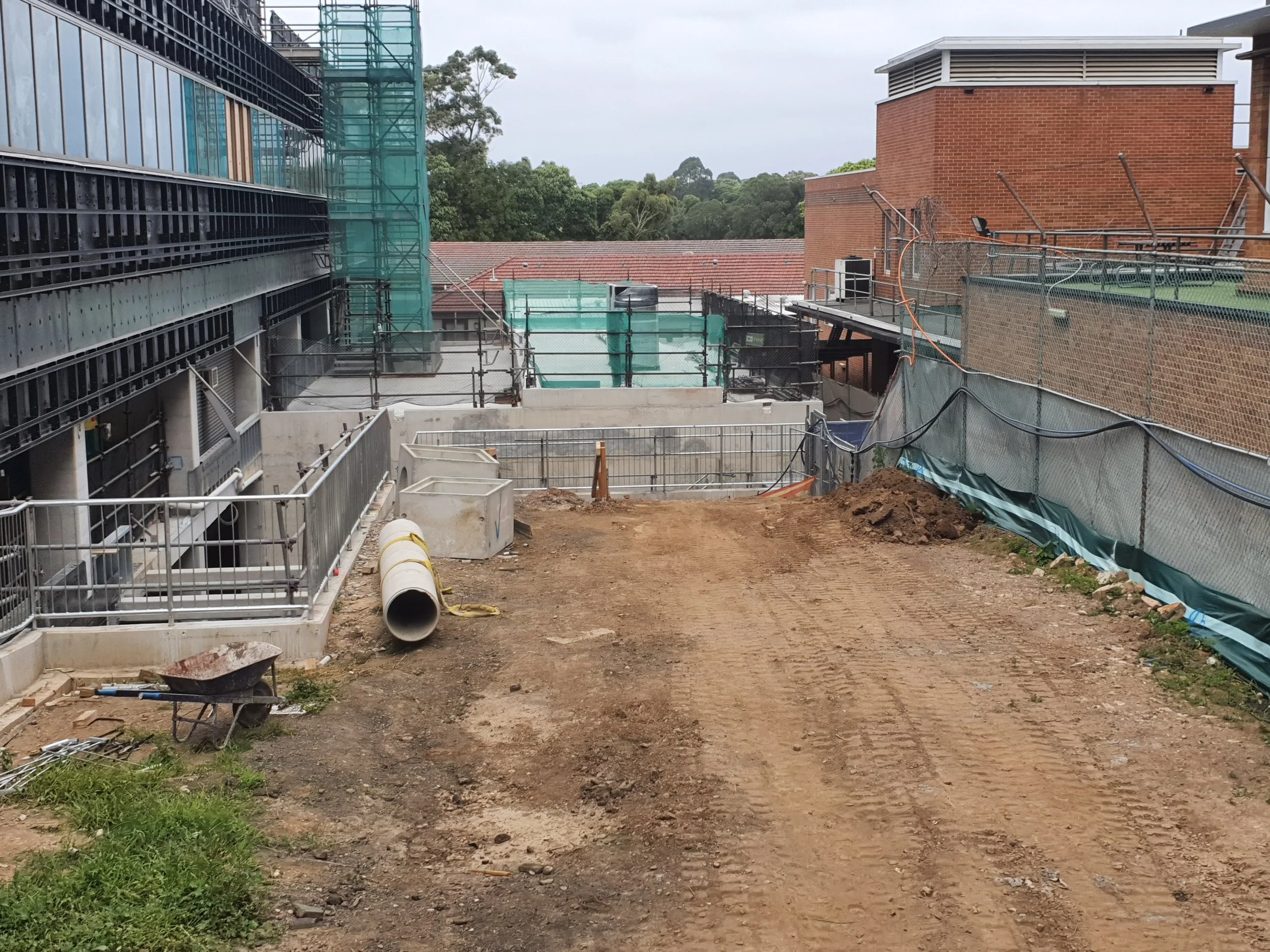 Construction site with dirt pathway, construction materials, safety barriers, and partially built structures, surrounded by trees and neighboring buildings.