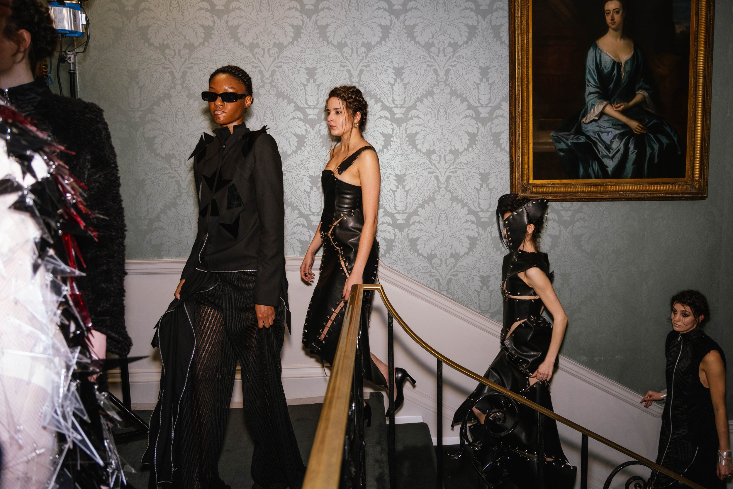 Models walking up the stairs at the Genaro Rivas Runway Show during London Fashion Week, black leather dresses, sunglasses