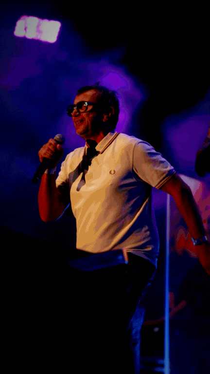Person performing with a microphone on stage, lit with purple and blue lights, wearing glasses and a white shirt.
