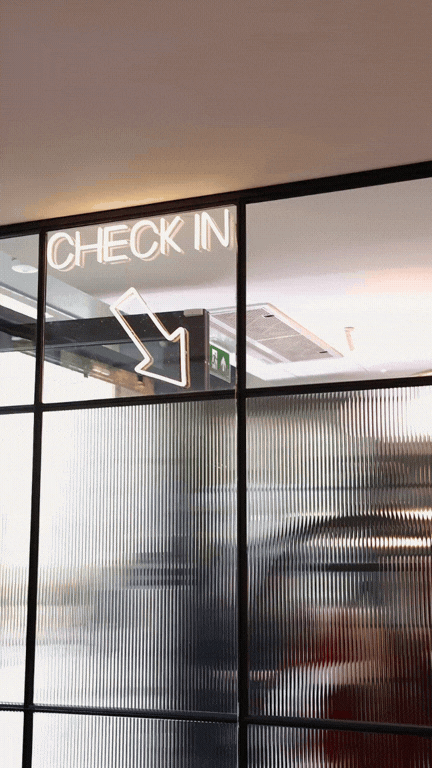 Neon sign that says 'CHECK IN' with an arrow pointing downward, located above a glass wall in an indoor setting.