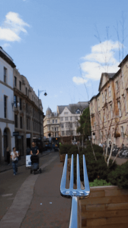 Close-up of a metal fork in the foreground on a city street with buildings, trees, and a clear blue sky.