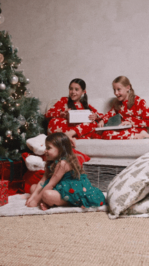 Three young girls in Christmas pajamas sitting near a decorated Christmas tree, opening presents indoors.