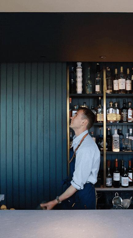 Bartender standing behind a bar with liquor bottles on shelves in the background, looking upward.