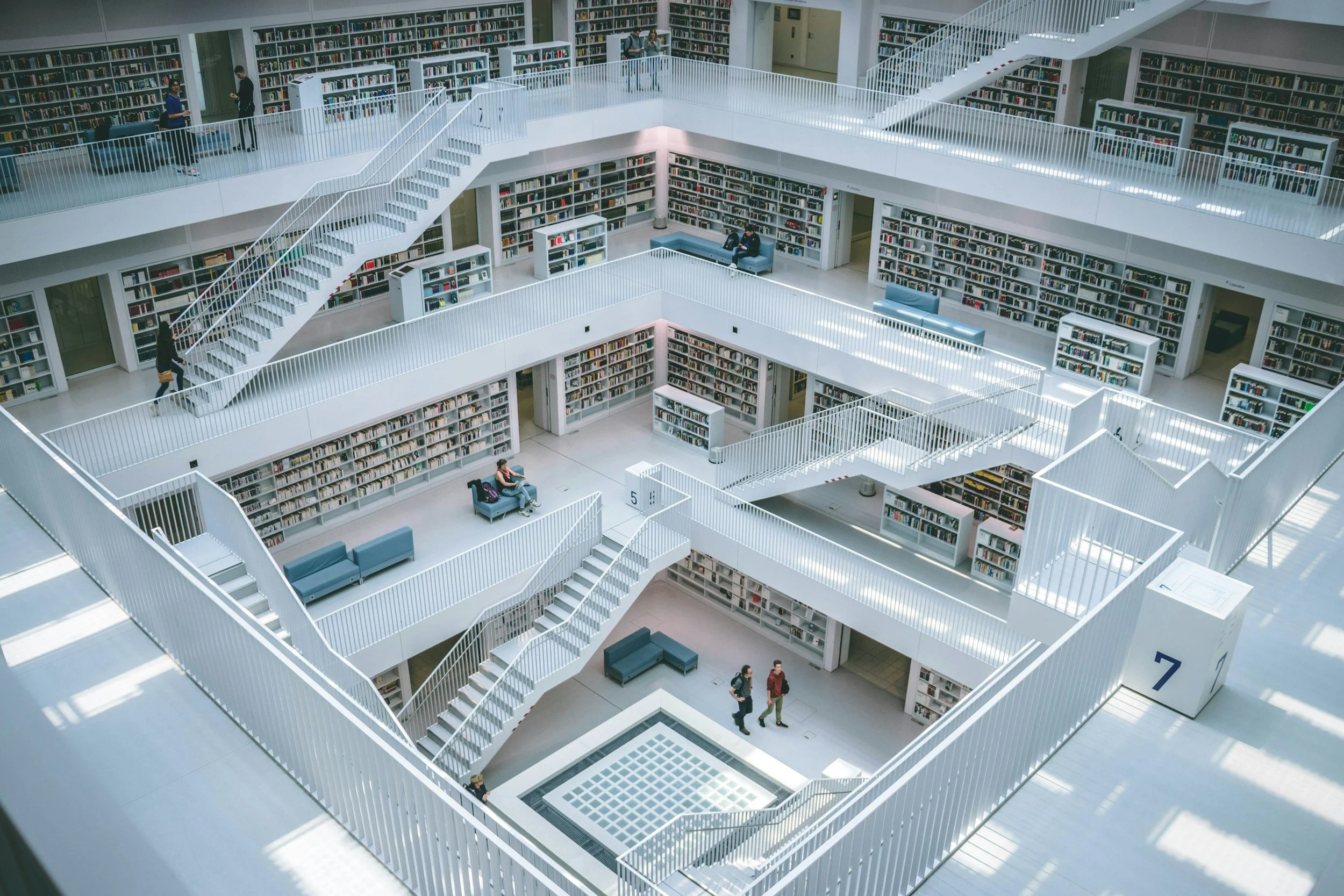 Modern library interior with multiple levels of bookshelves, white walls, staircases, and people reading or walking.
