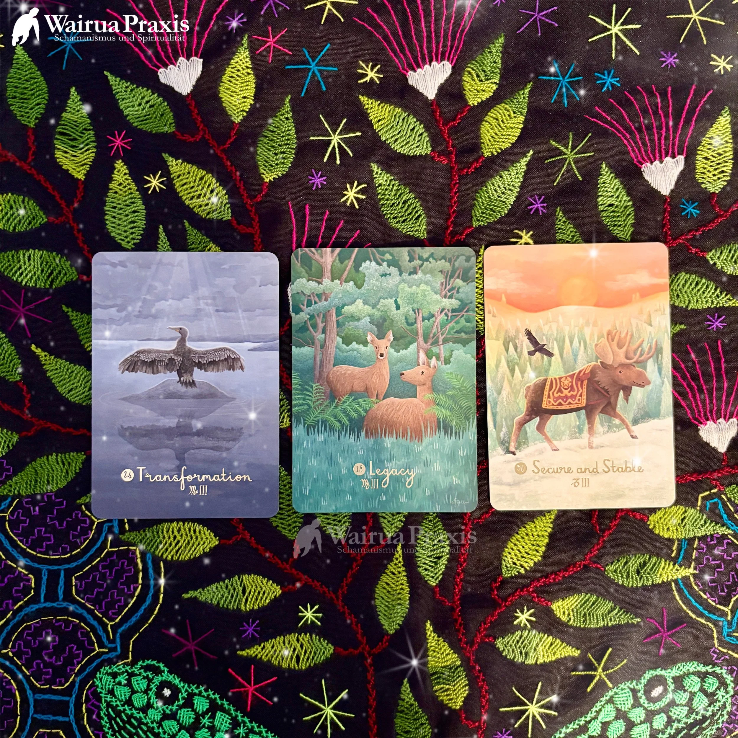 Drei Tarot-Karten auf einer schwarzen, bestickten Stoffdecke mit bunten, blattförmigen und sternförmigen Mustern. Die erste Karte zeigt einen schottischen Kranich mit dem Wort "Transformation". Die zweite Karte zeigt Rehe in einem Wald mit dem Wort "