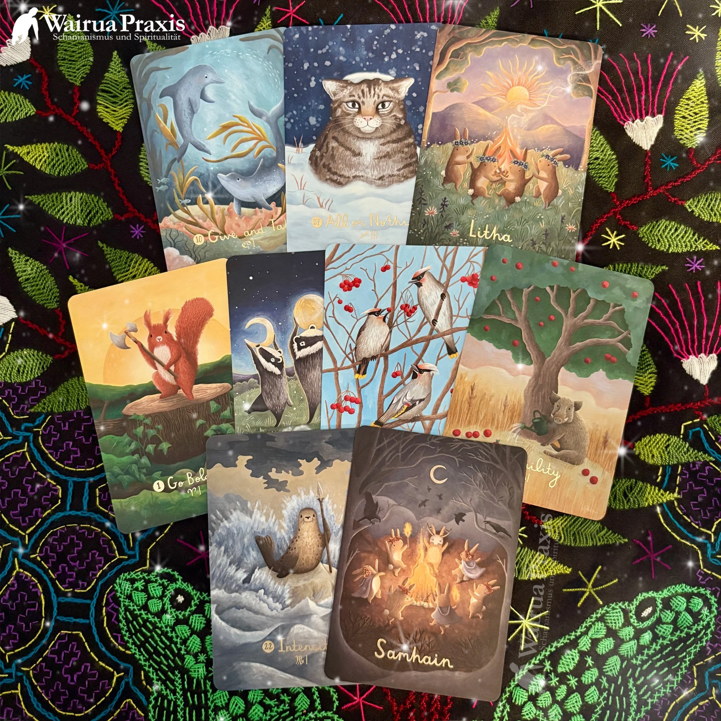 Eine Vielzahl von Tarotkarten mit Tier- und Naturmotiven liegt auf einem schwarzen Tuch mit bunten, floralen Stickereien.