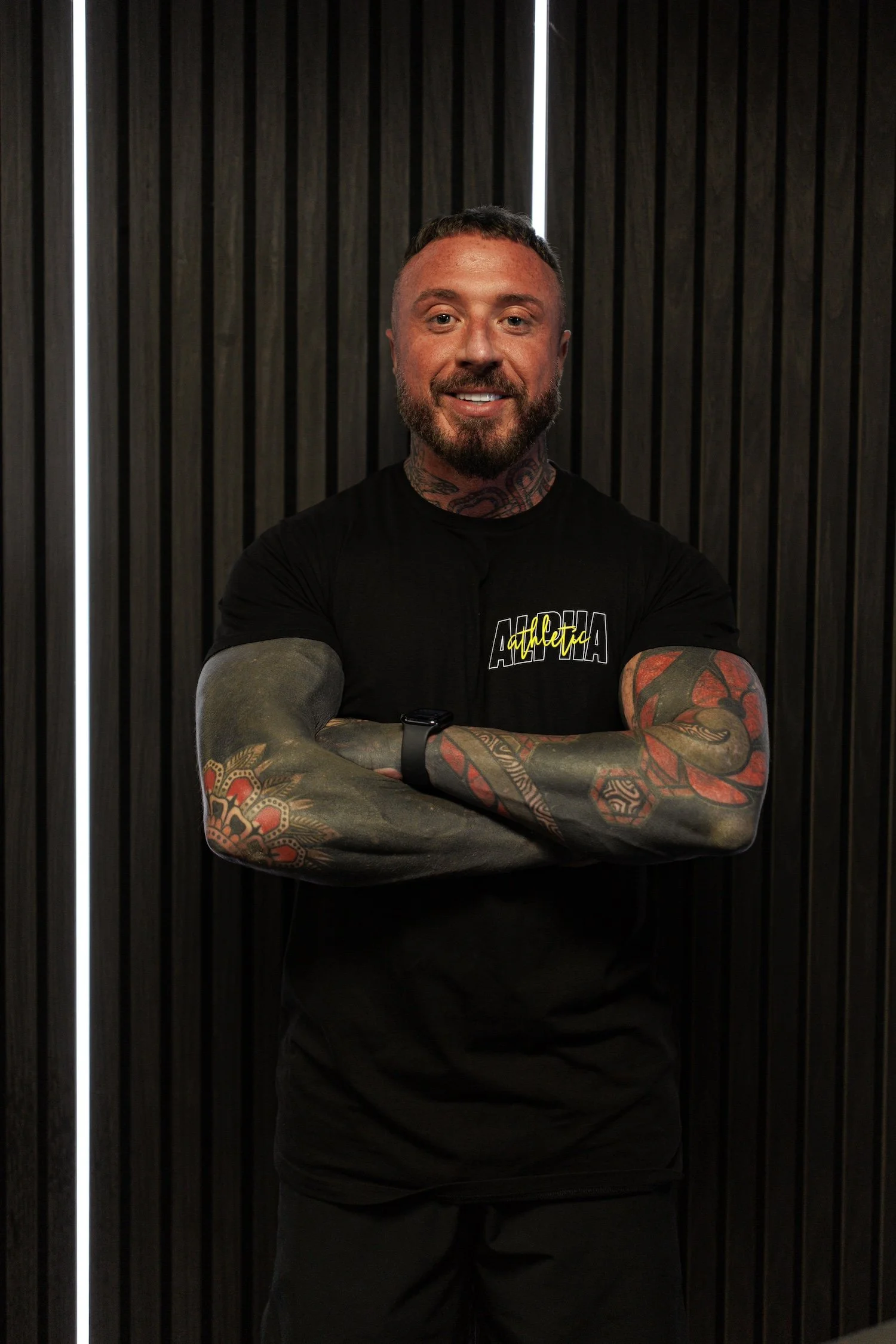 A male personal trainer is standing with his arms crossed and smiling in an Alpha Athletic T Shirt.