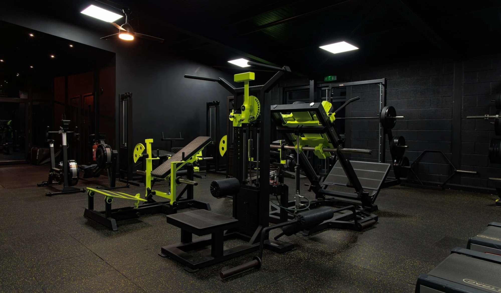 Empty gym with various black and green workout equipment, black walls, and ceiling, bright ceiling lights, rubber flooring.