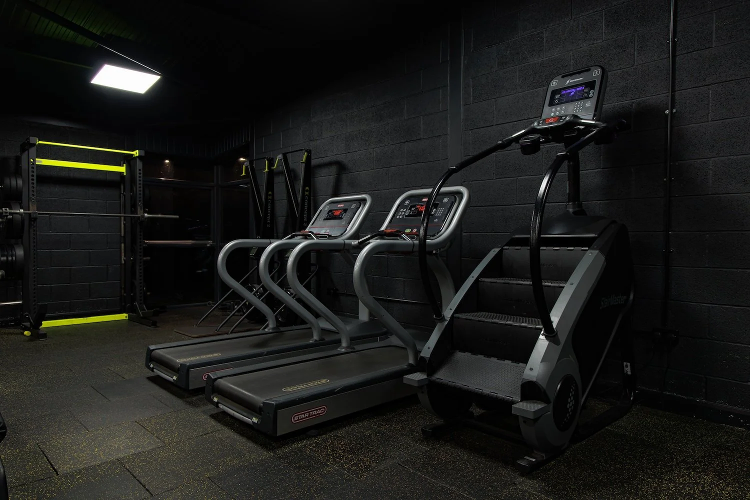 A gym room with a treadmill, elliptical trainer, and stair climber in front of a black brick wall.