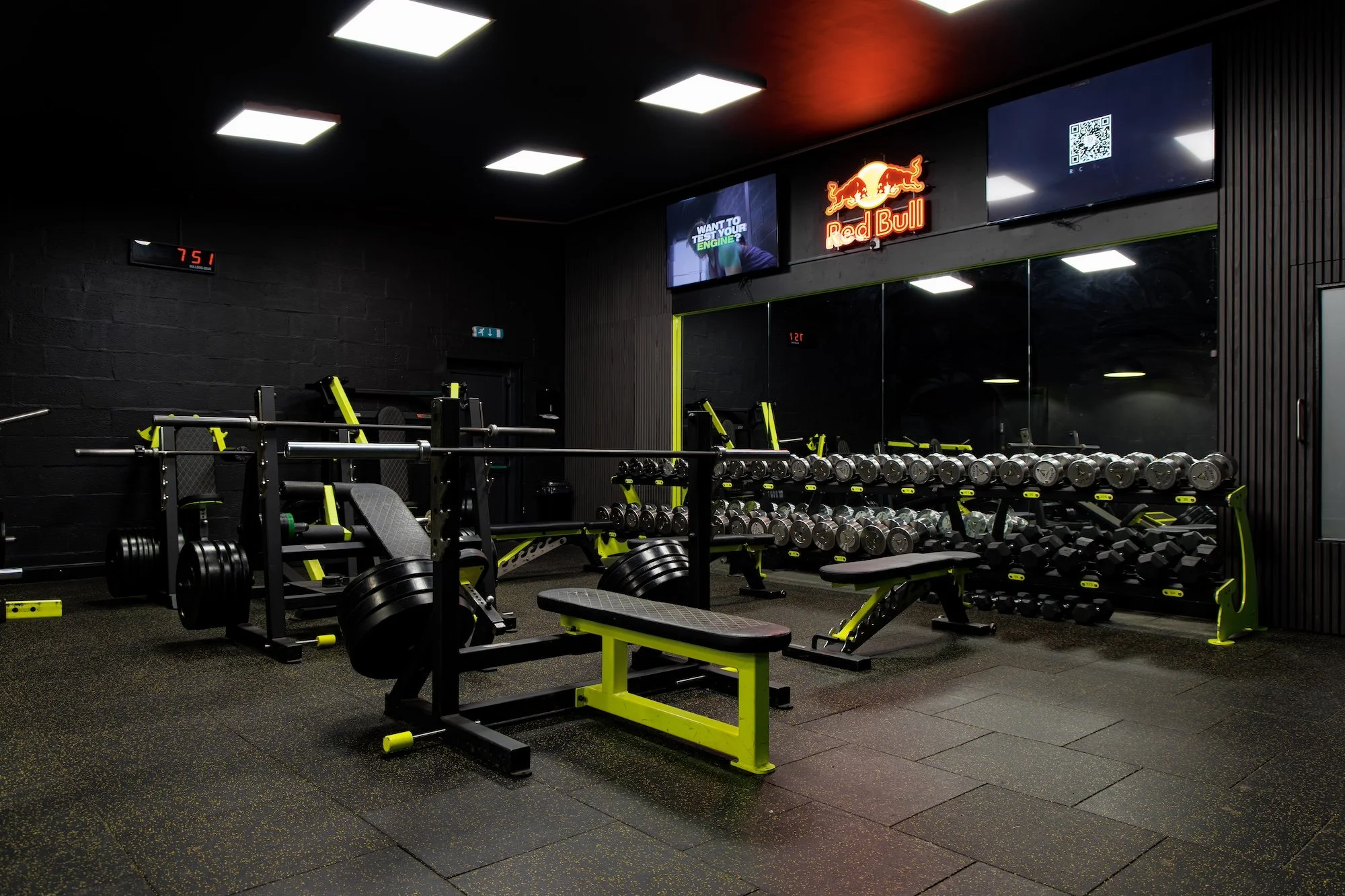A gym with weightlifting equipment and mirrors, illuminated by ceiling lights, including benches, dumbbells, and a rack of weights, with digital displays and neon Red Bull sign on the wall.