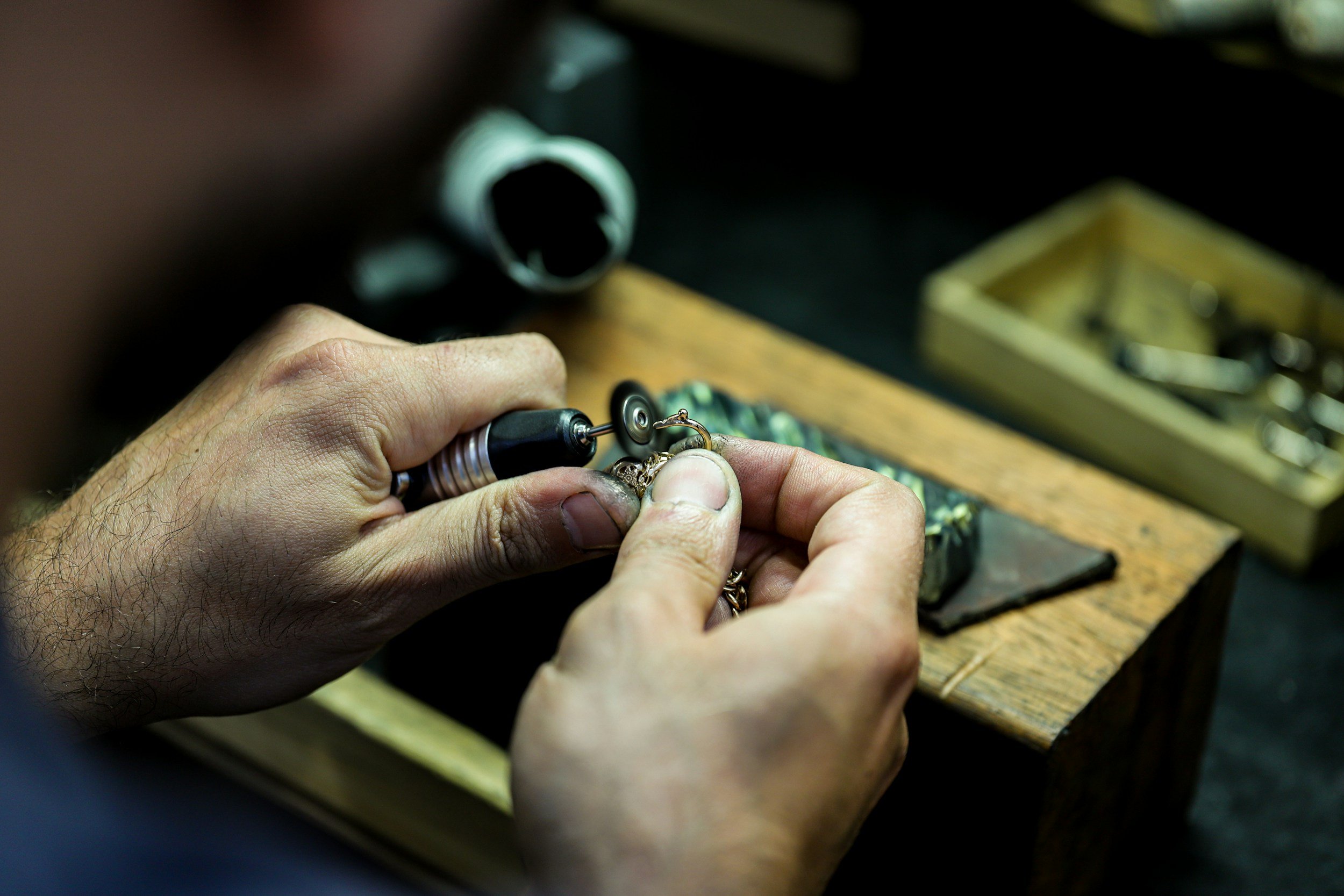 A person using a rotary tool to engrave or work on a piece of jewelry, with a wooden work surface and a small wooden box containing jewelry-making tools in the background.