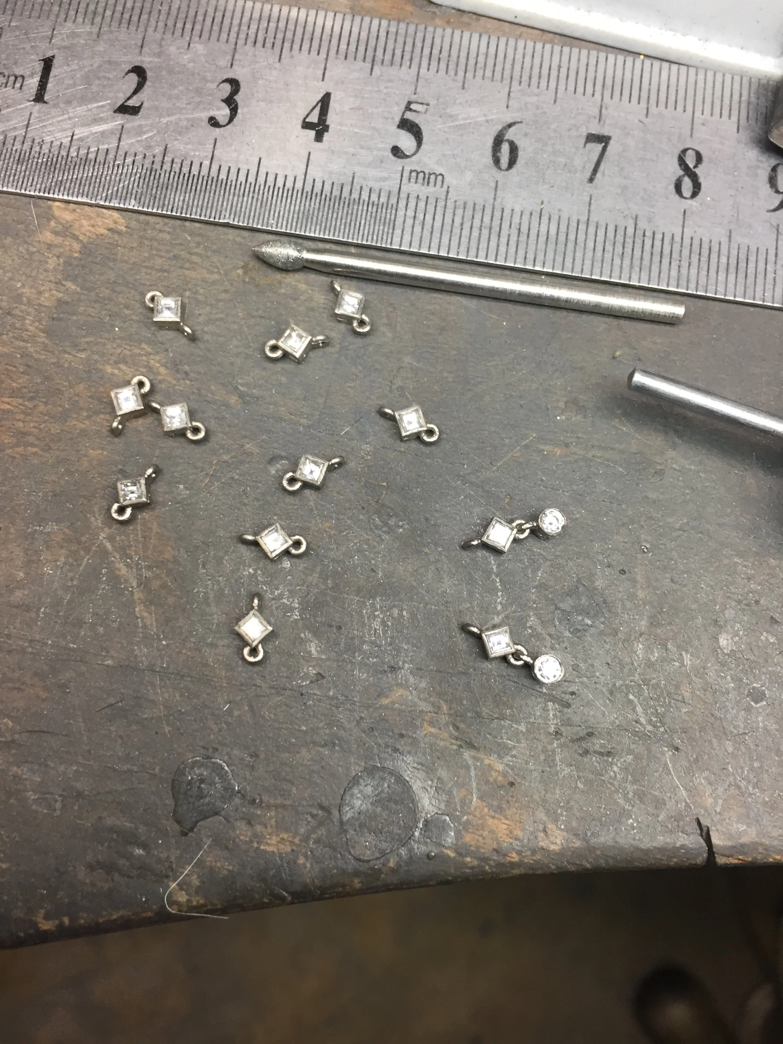 Small metal jewelry components with square and round clear stones, scattered on a workbench next to a metal ruler with millimeter markings and metal tools.