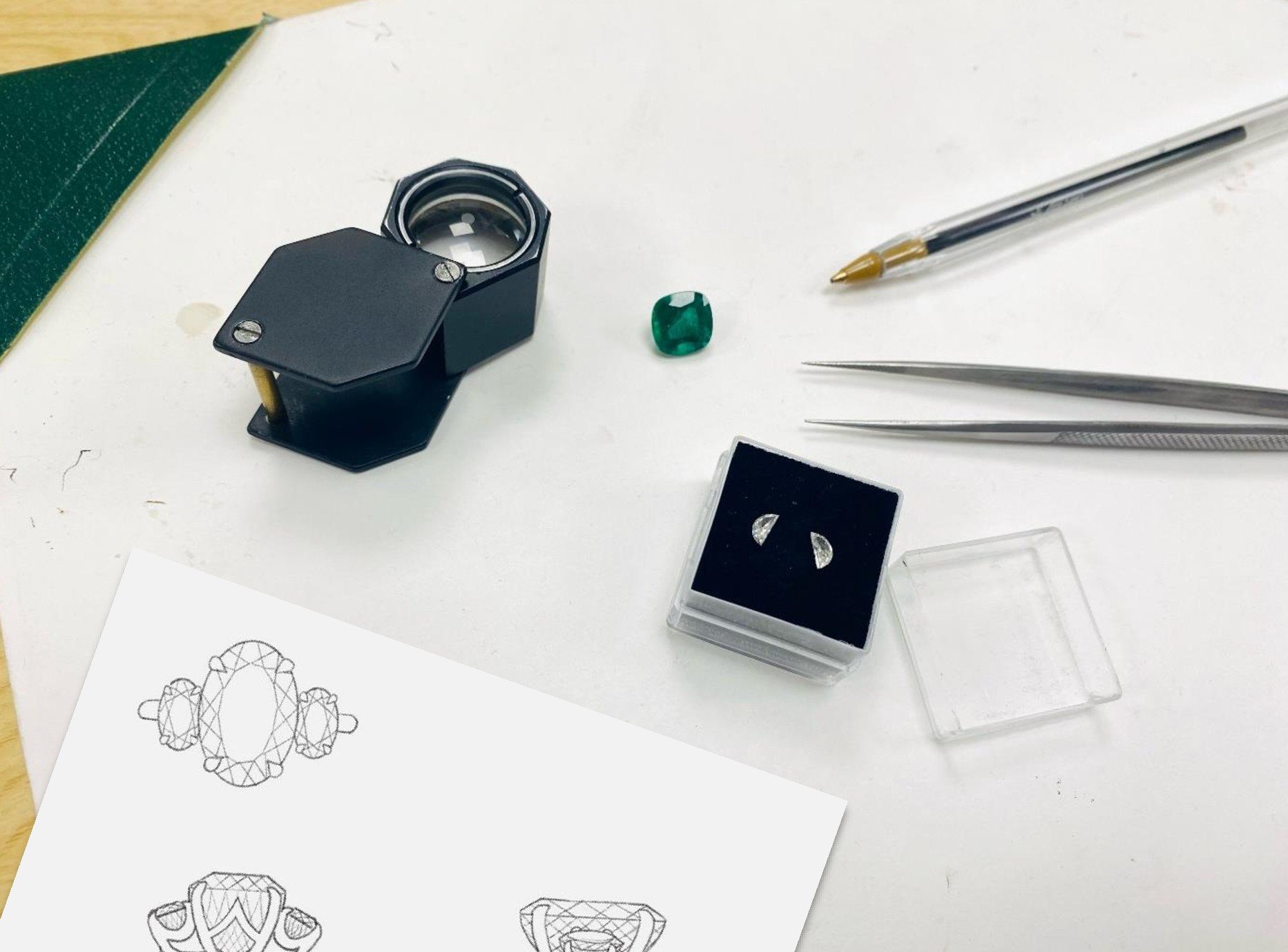 Jewelry design tools including a watch movement part, a small green gemstone, pair of diamond earrings, tweezers, precision tools, a sketch of jewelry designs, and a black display box on a white surface.