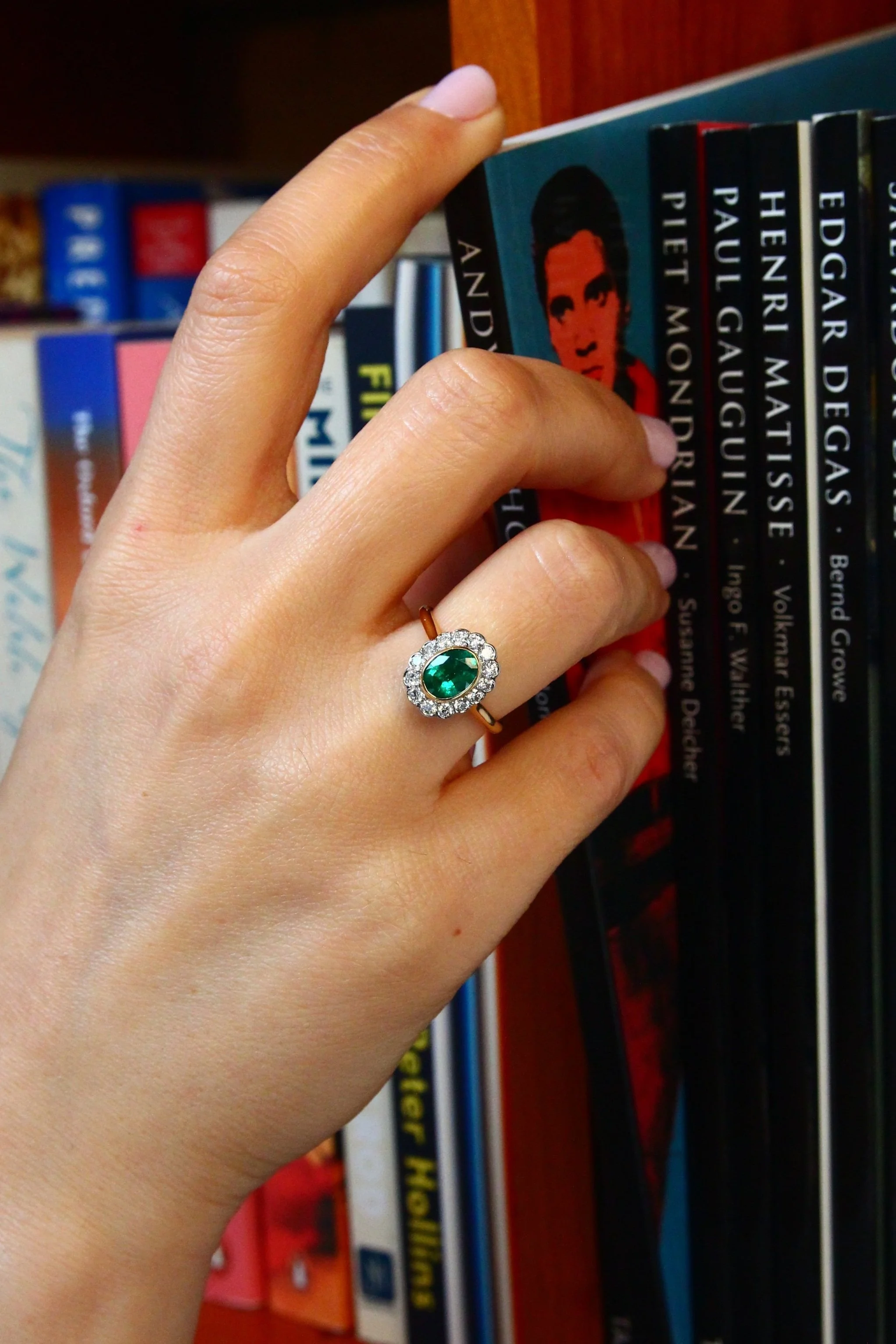 A hand with a ring featuring a large green gemstone surrounded by small clear stones, holding a book on a shelf filled with various books.