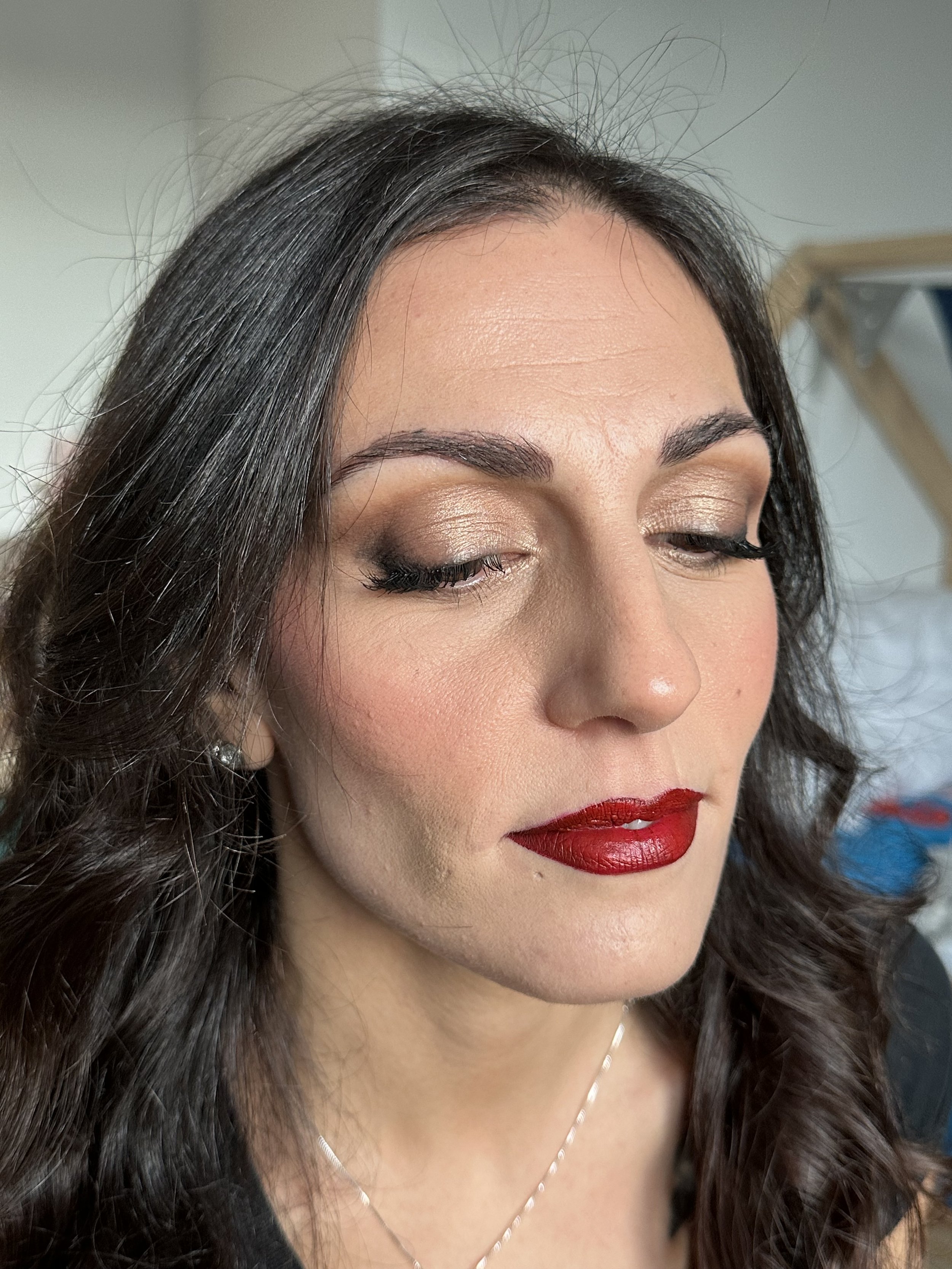 Donna con rossetto rosso intenso, occhi chiusi e trucco marrone dorato, capelli scuri ondulati.
