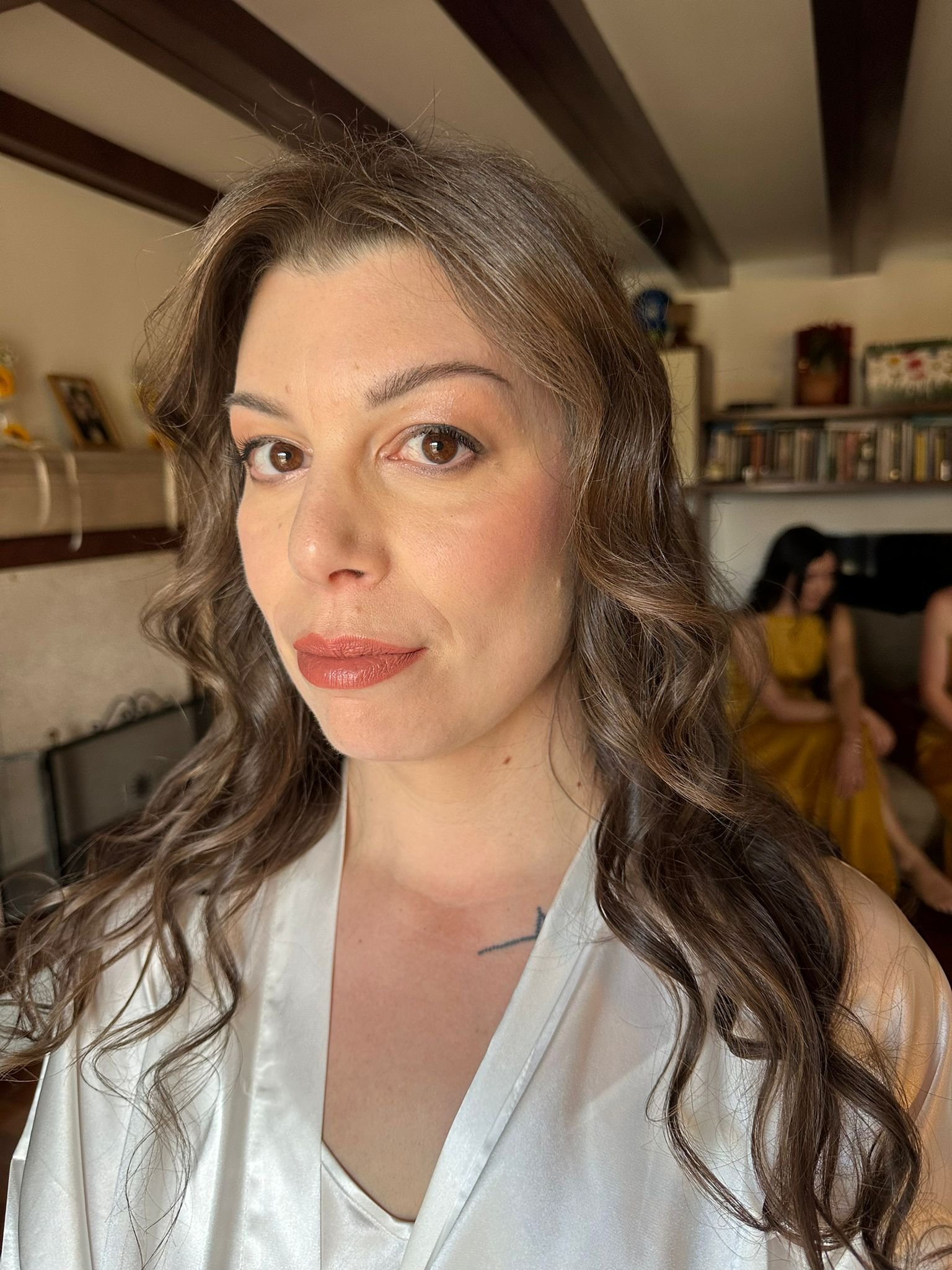 Donna con capelli lunghi e ricci, indossa una vestaglia bianca, in una stanza con scaffali sullo sfondo.