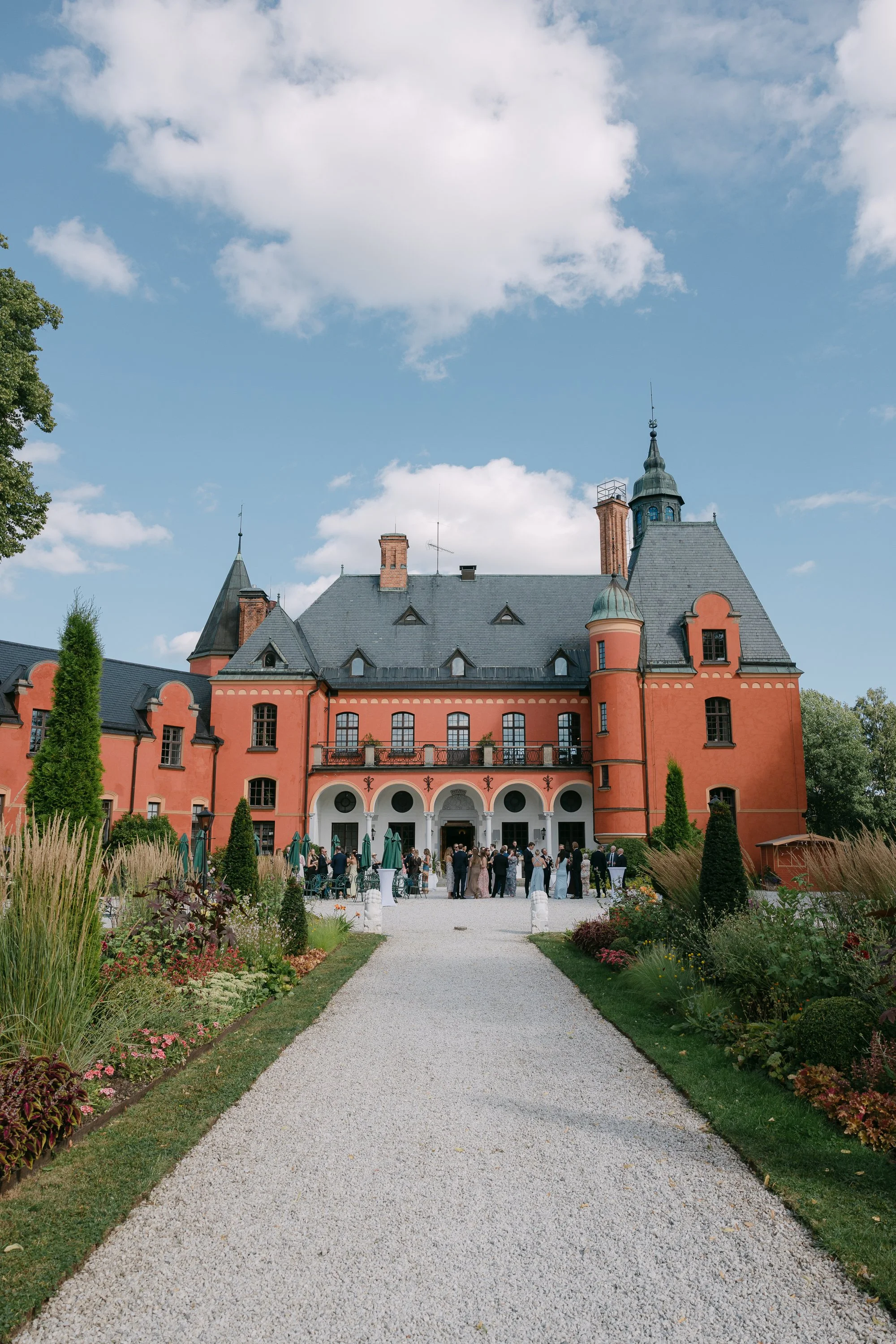 Ett slottliknande byggnad i rosa färg med många torn och turrets, omgivet av en trädgård med blomsterbäddar och en gångväg som leder till entrén. Det är ett evenemang pågår utanför där människor samlas, med gröna parasoller och utomhusmöbler.