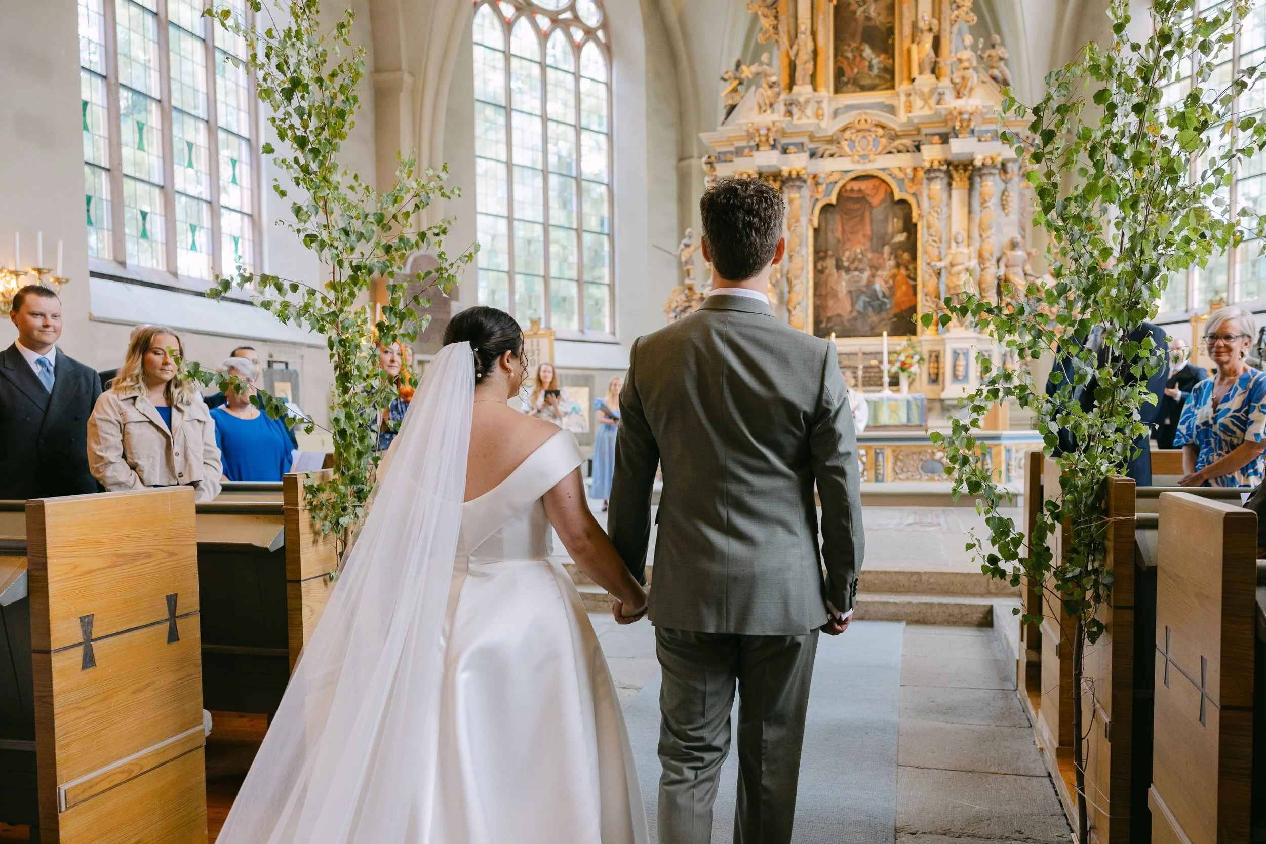 En brud och en brudgum står tillsammans under ett bröllopsceremoni i en kyrka, omgivna av gäster och dekorationer.