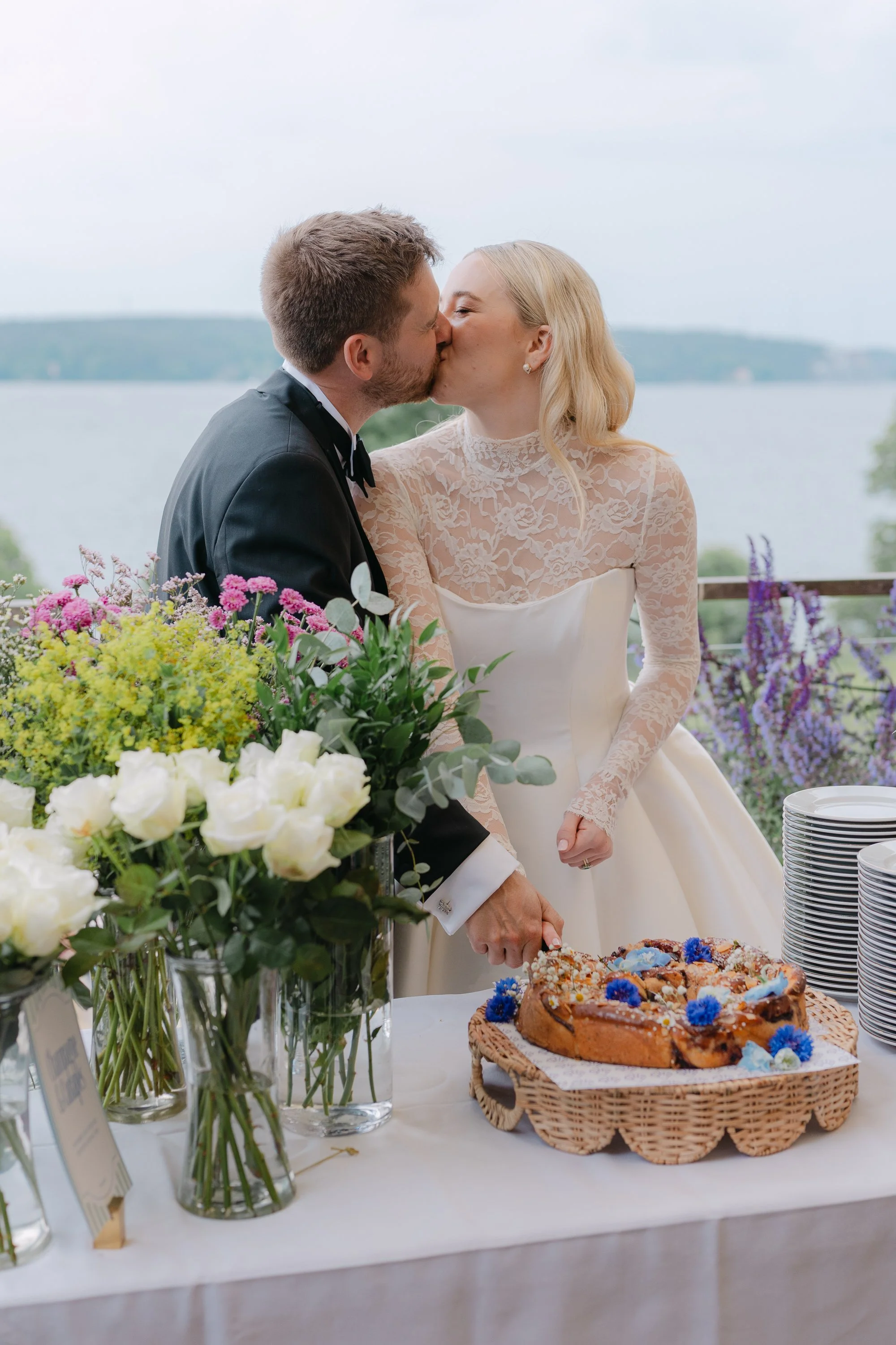 Ett par kysser vid ett bröllopsfirande, med en duk av blommor och tårtor på bordet, utomhus och med en sjö i bakgrunden.