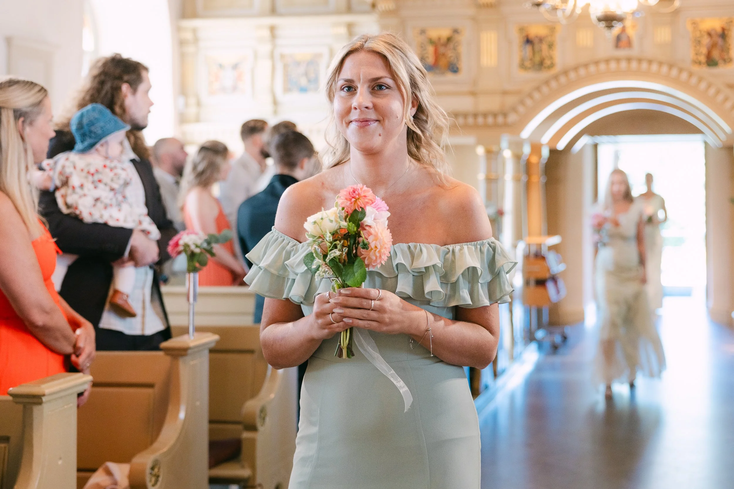 En glädjefull kvinna i en ljusgrå klänning, brudtärna, håller en bukett blommor, står i en kyrka under ett bröllop.   Av Sofia Wilhelmina bröllopsfotograf i Stockholm.