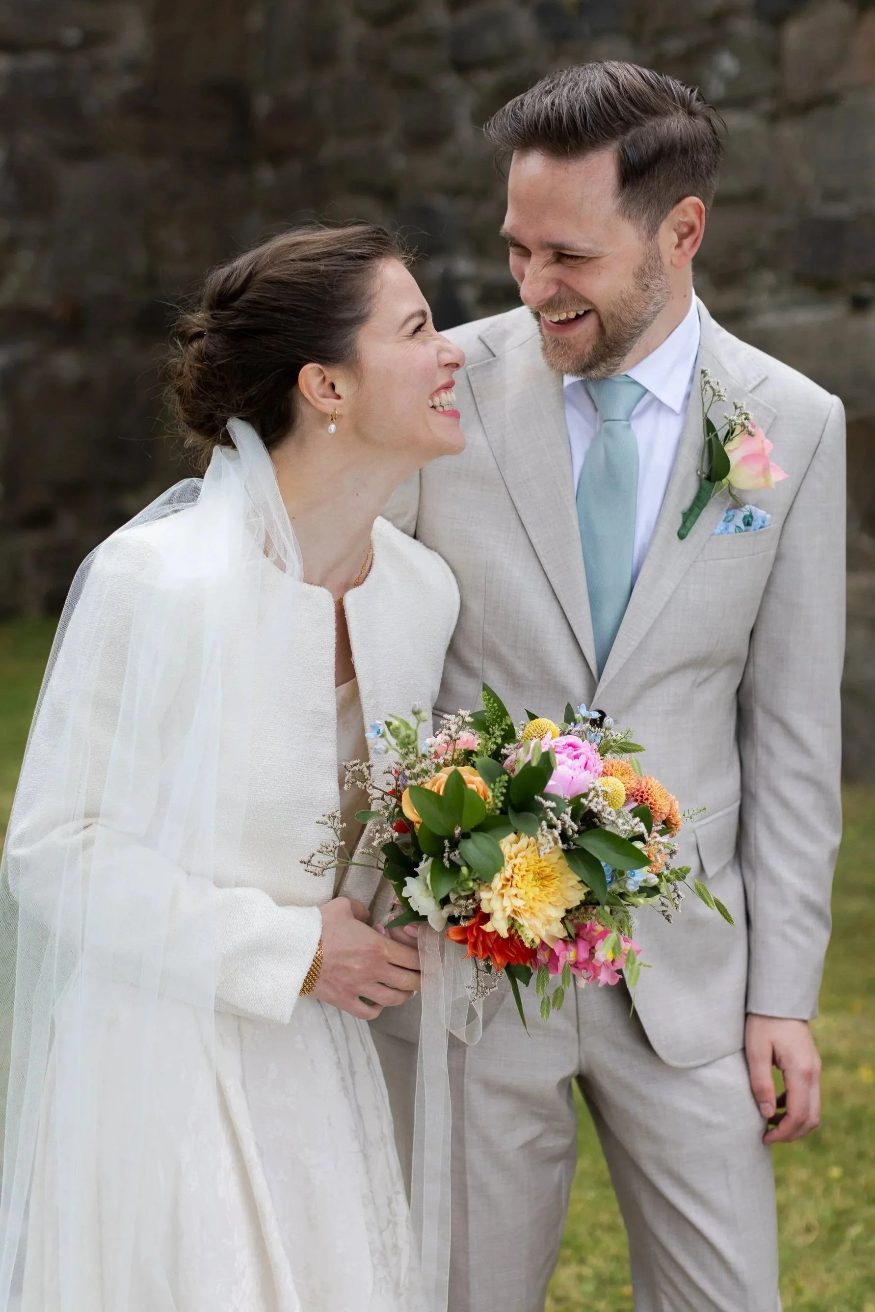 Brud och brudgum står i naturen, skrattar för fullt och ser på varandra