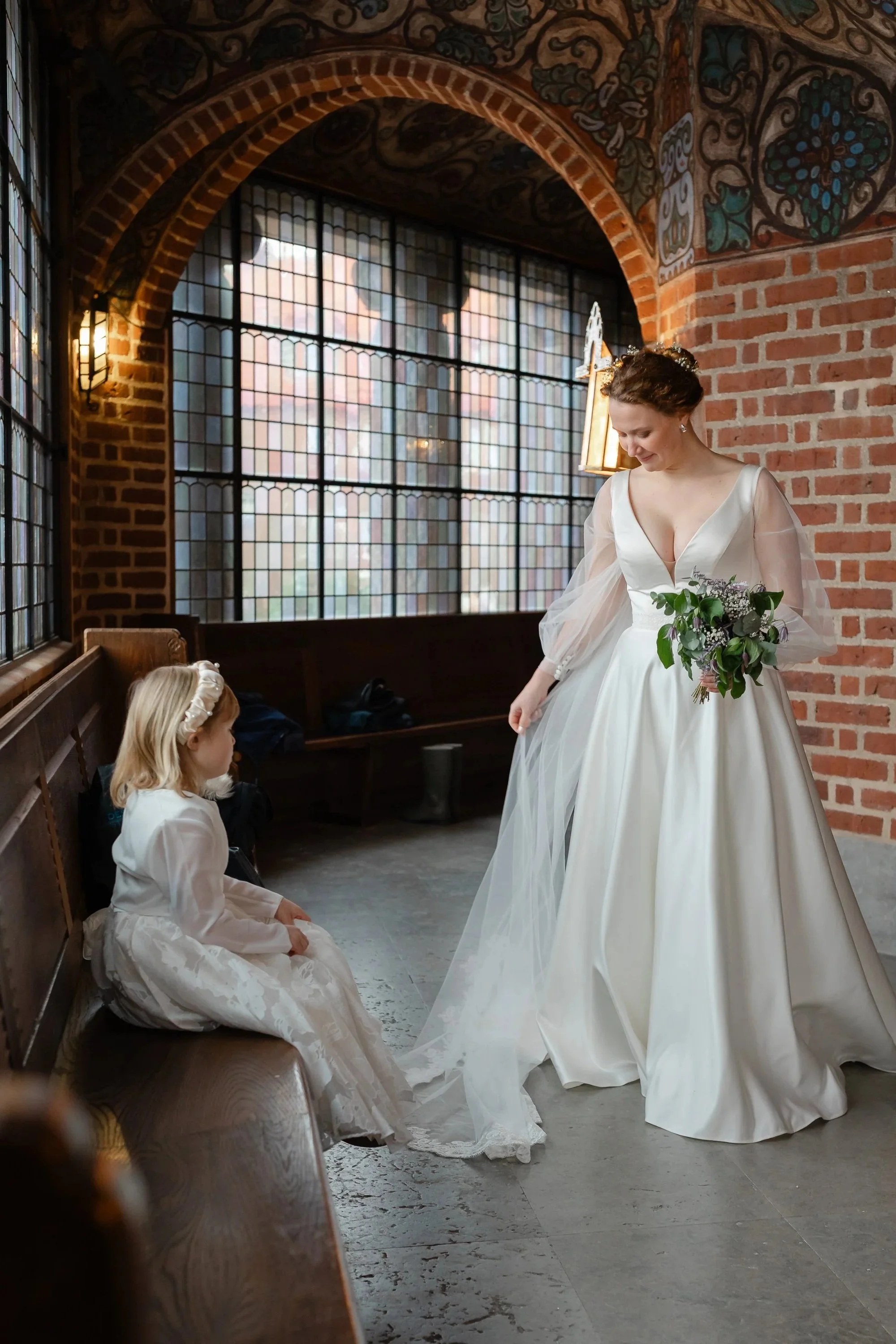 En brud i vit brudklänning och ett litet barn i vit klänning och hårband sitter på en bänk i ett rum med tegelväggar och stora fönster, i Engelbrektskyrkan i Stockholm.