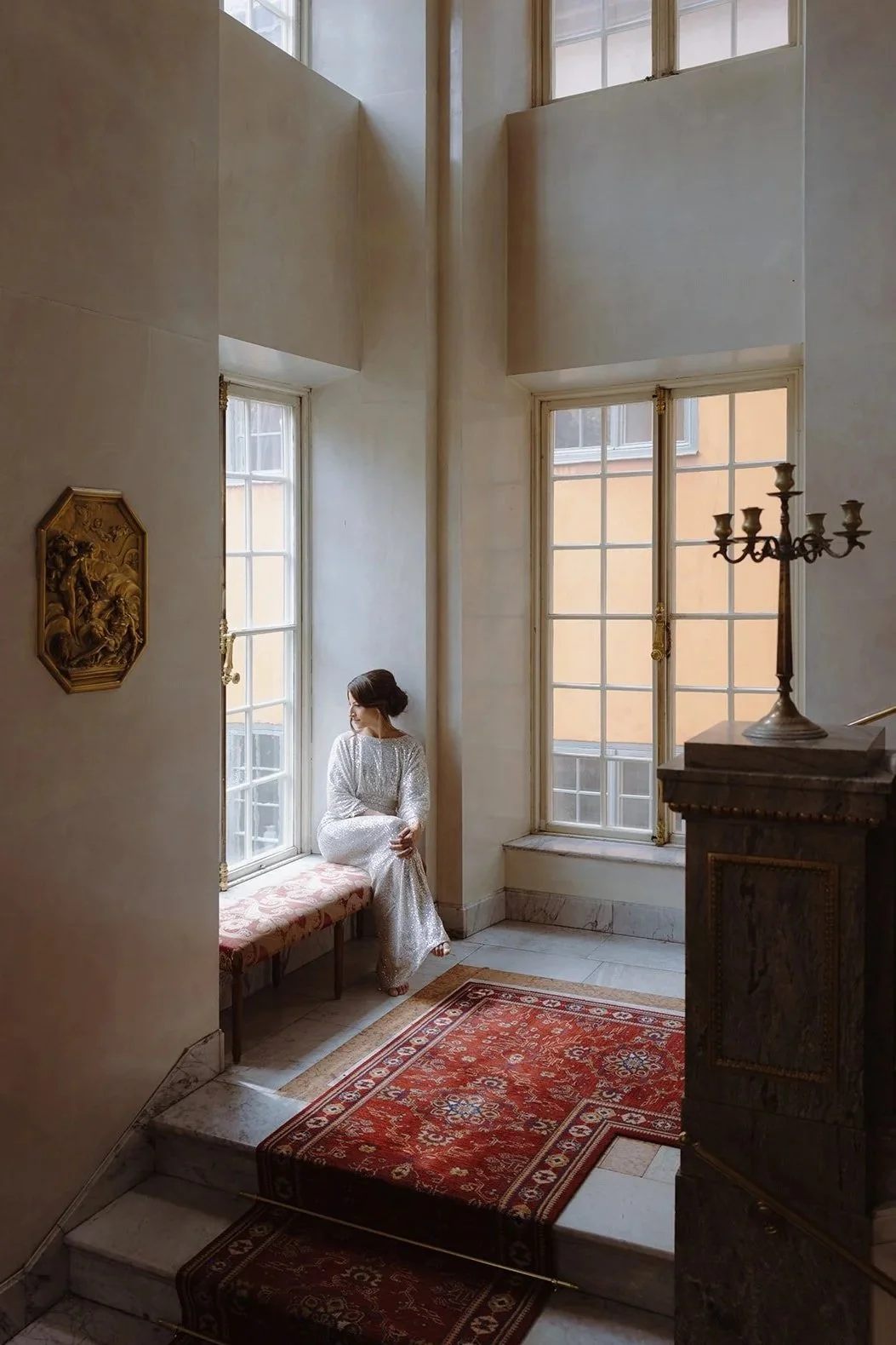 En kvinna i en vit klänning sitter på en bänk vid ett stort fönster i ett ljust rum.