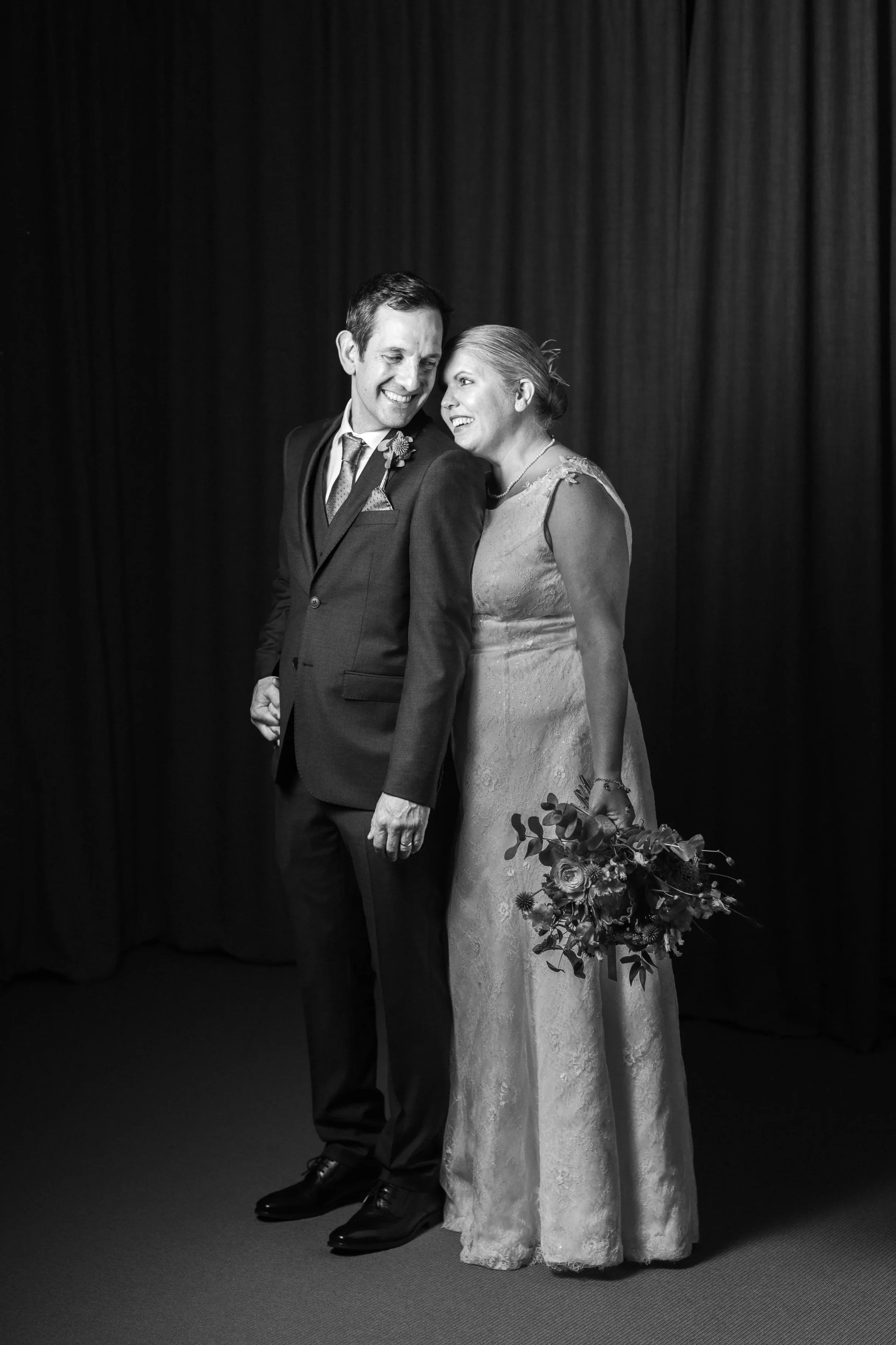 Ett svartvitt foto av två personer på bröllop, en man och en kvinna, som ler mot varandra framför en mörk bakgrund.