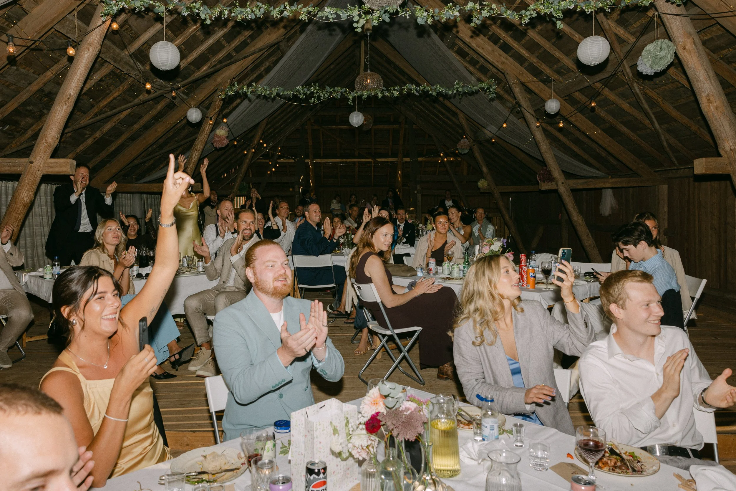 En grupp människor i festkläder som skrattar och applåderar inomhus under en fest i ett loft med träbalkar och dekorationer, inklusive papperslyktor och blommor.  Av Sofia Wilhelmina bröllopsfotograf i Stockholm.