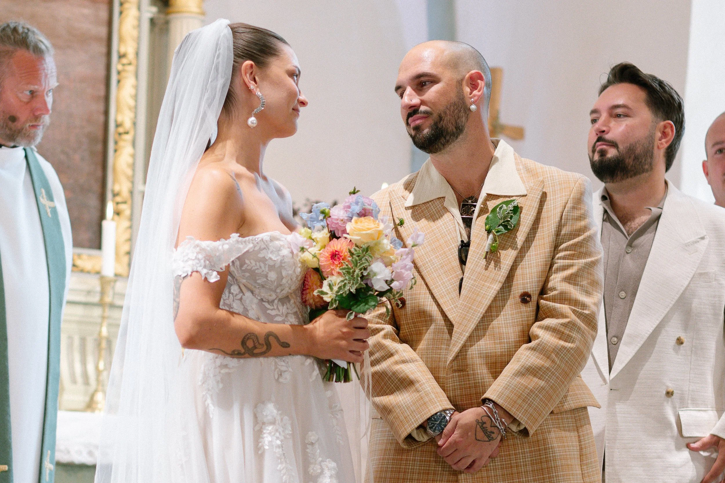 En brud och en brudgum står framför en kyrka under ett bröllop, bruden håller en bukett blommor och de ser varandra i ögonen, personen i förgrunden till vänster är en präst.  Av Sofia Wilhelmina bröllopsfotograf i Stockholm.