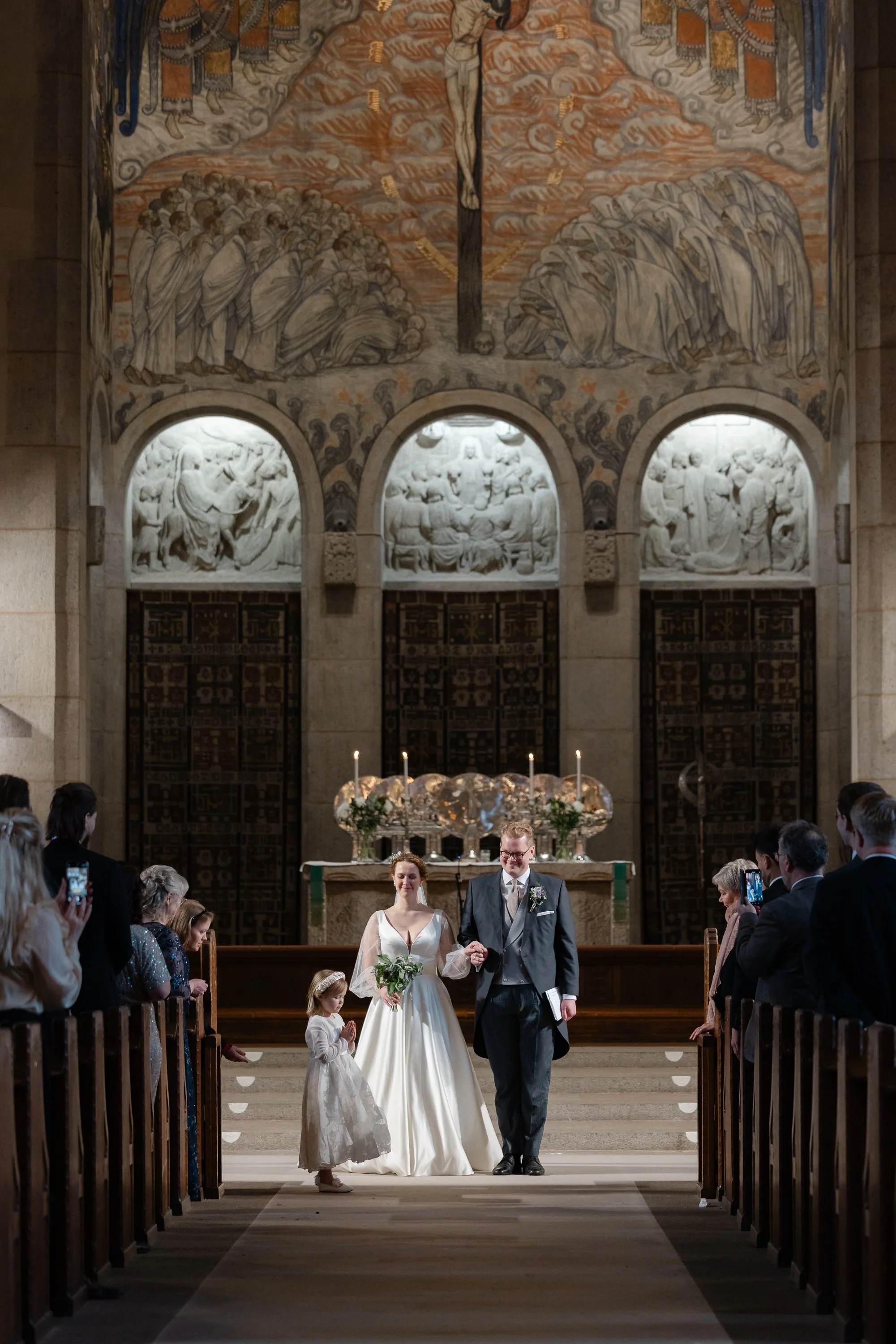 Bröllopsceremoni i en kyrka med bruden, brudgummen och ett ungdjur som håller handen, omgiven av gäster.