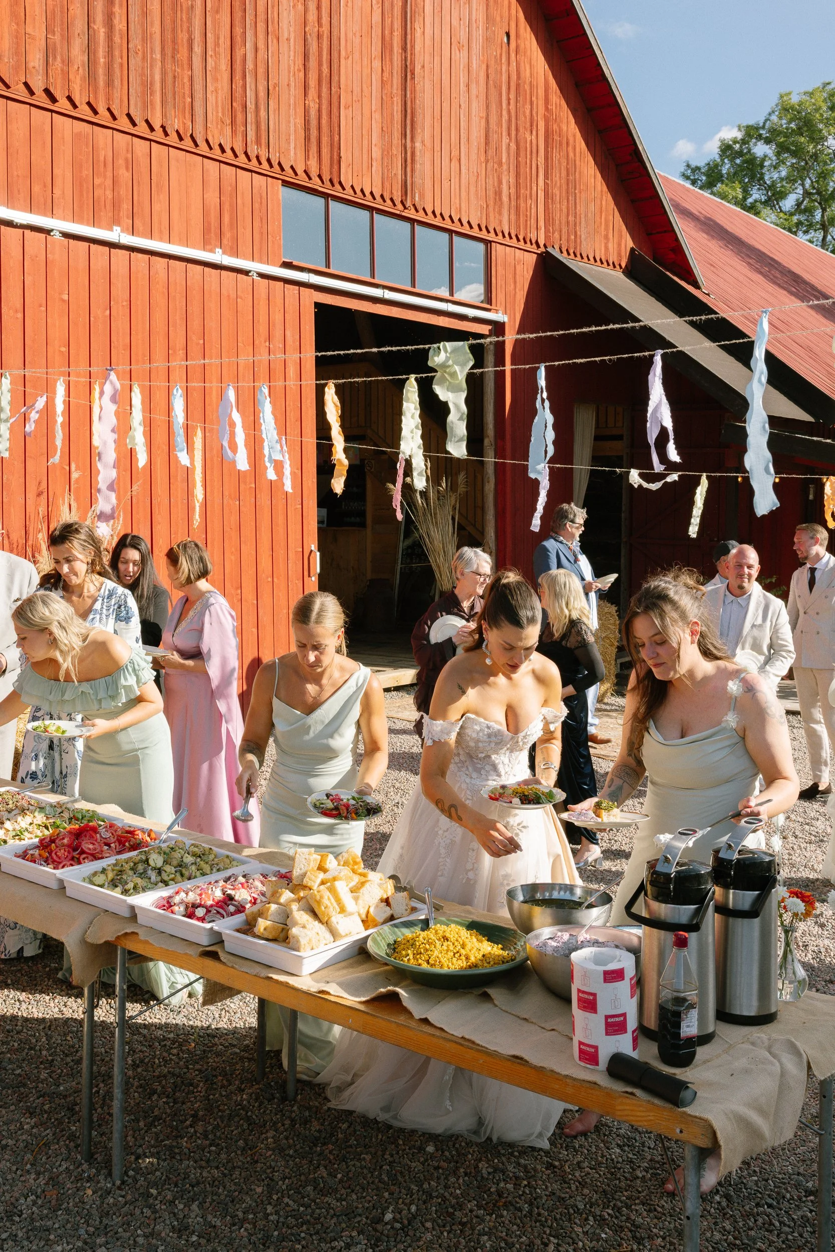 Bröllopsgäster serverar mat utomhus framför ett rött trähus, med dekorativ tygremsa hängande från linnet.