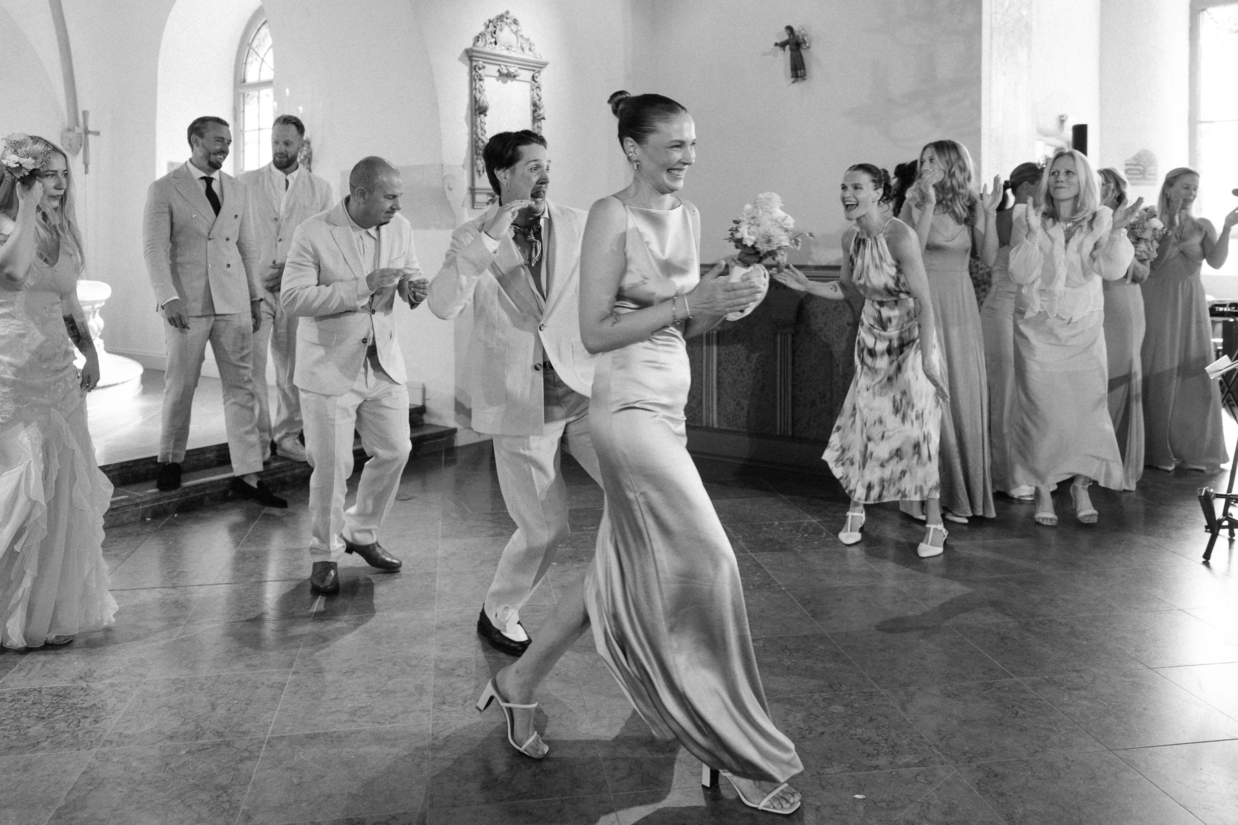 Svartvitt foto av ett bröllop, där bruden och gästerna dansar och har roligt inomhus.