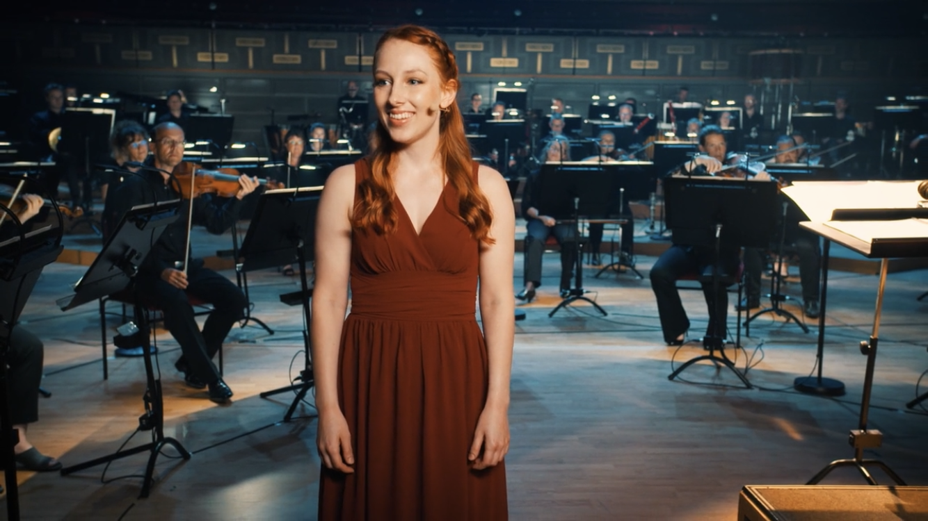 En ung kvinna i röd klänning klädd står framför en orkester i en konsertsal.