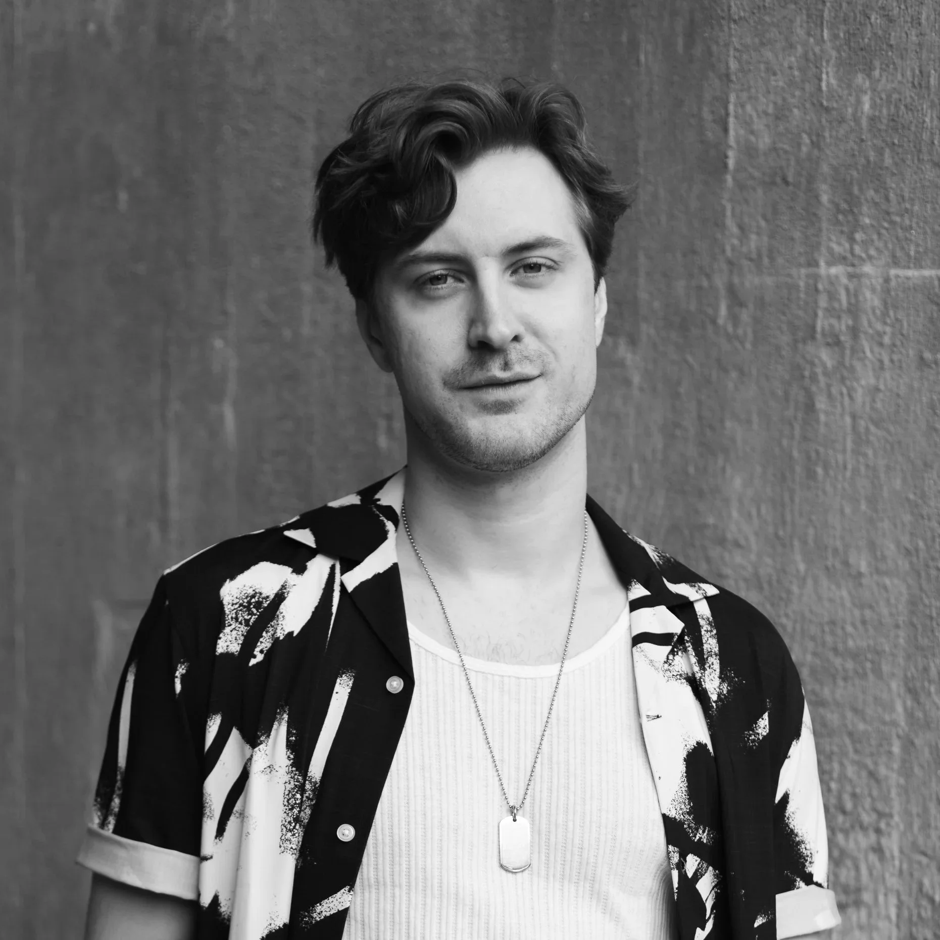 Svartvit porträttfoto av en ung man med lockigt hår, lätt leende, iförd en vit t-shirt och en mönstrad öppen skjorta, stående mot en vägg.