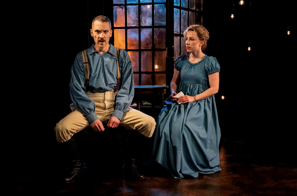 Två personer på scen, en man och en kvinna, i ett teaterspel. Mannen sitter på en stol, woman står bredvid, båda i tidsenliga kläder.