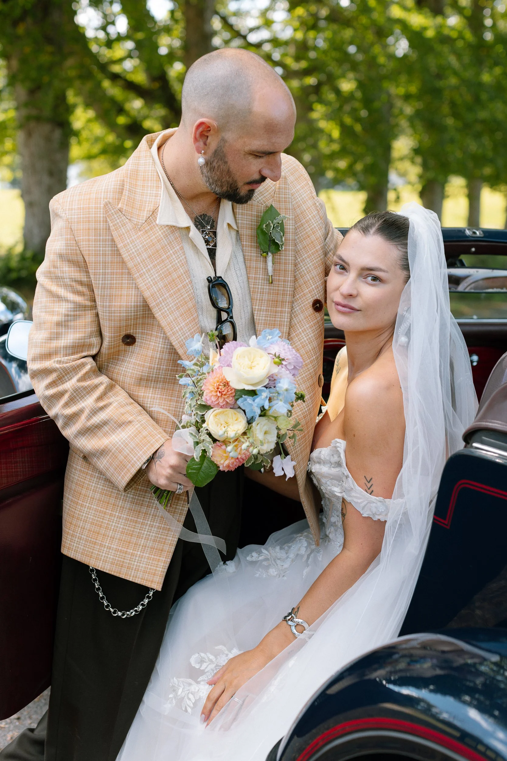En man och en kvinna på bröllop, med mannen som håller en bukett blommor, står nära en öppen bil, med träd och grönt bladverk i bakgrunden.  Av Sofia Wilhelmina bröllopsfotograf i Stockholm.