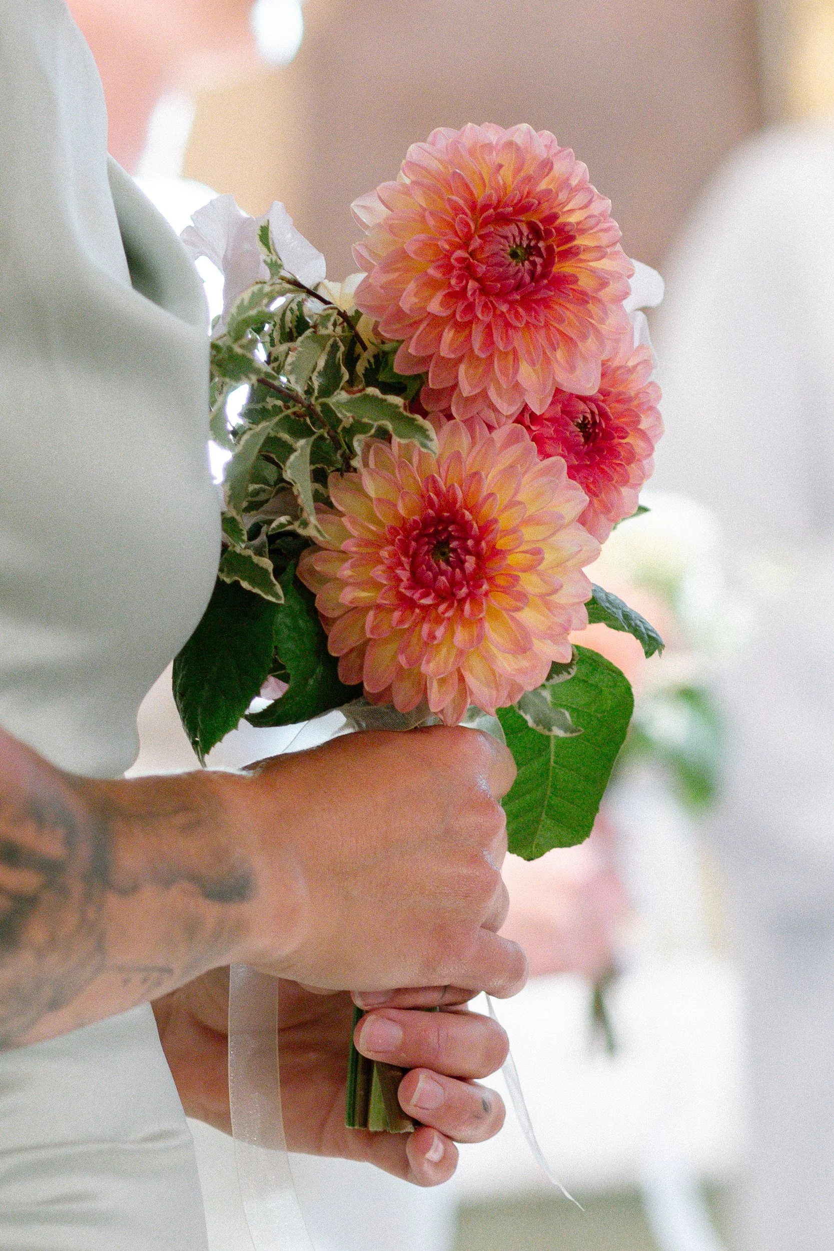 En person håller i en bukett med tre rosa och gula blommor och gröna blad, och blicken är nedåt.  Av Sofia Wilhelmina bröllopsfotograf i Stockholm.