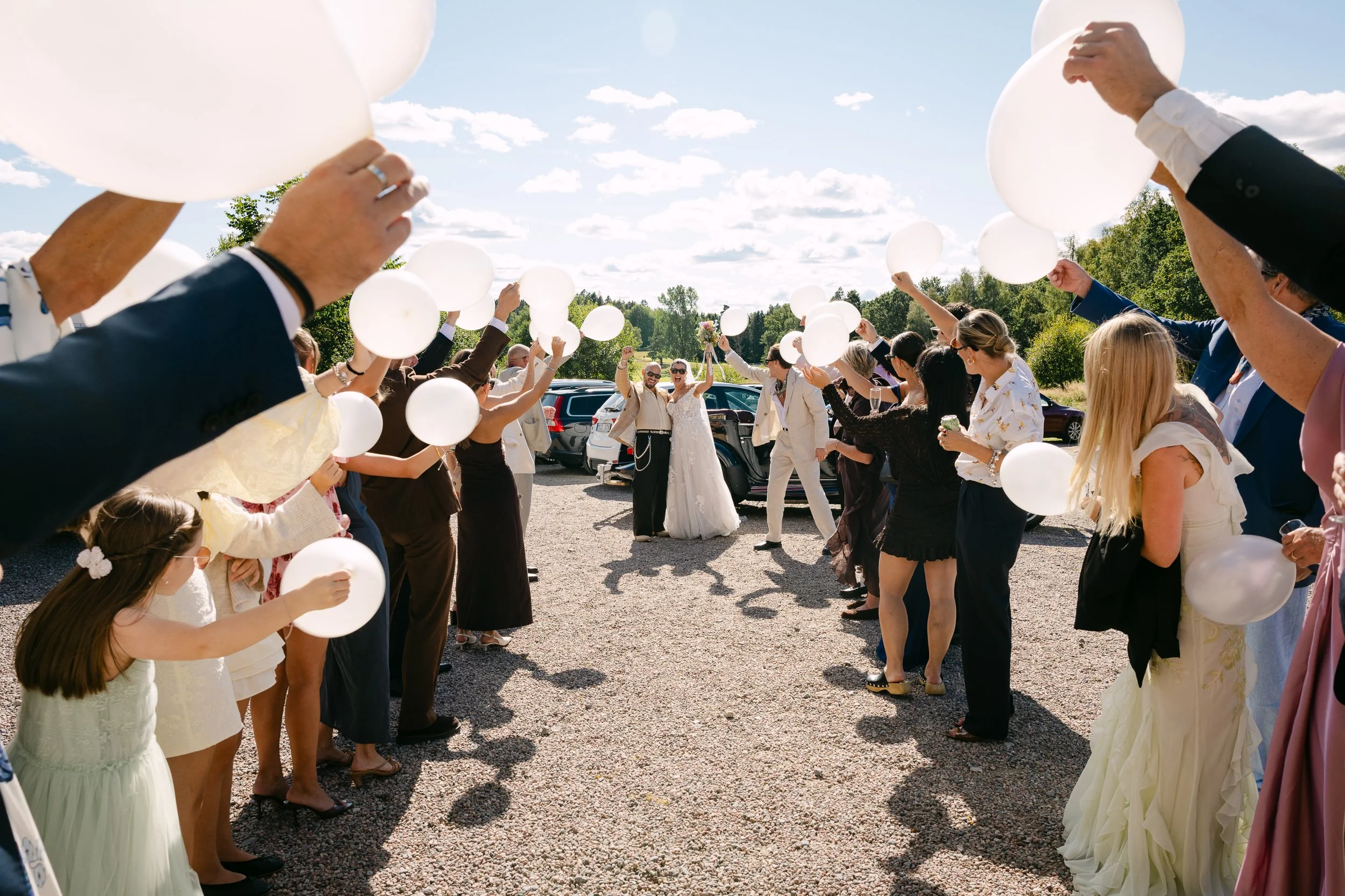 Flera personer samlade för ett bröllop, håller vita ballonger i handen, står i två rader under en klar himmel, med natur och bilar i bakgrunden.