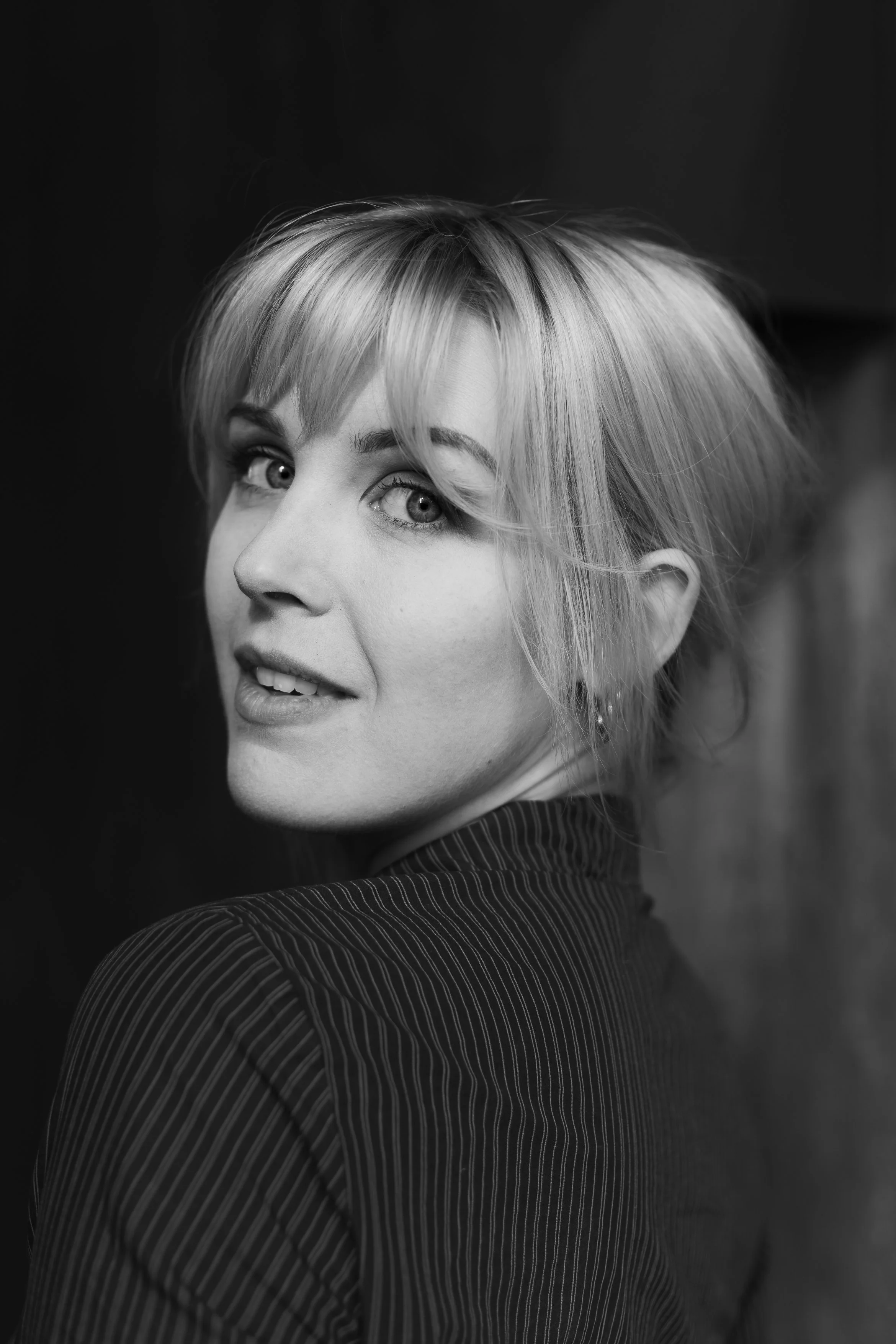 Svartvit porträttbild av en blond kvinna med löst, lite rufsigt hår, som tittar över axeln mot kameran, med en lätt leende ansiktsuttryck.