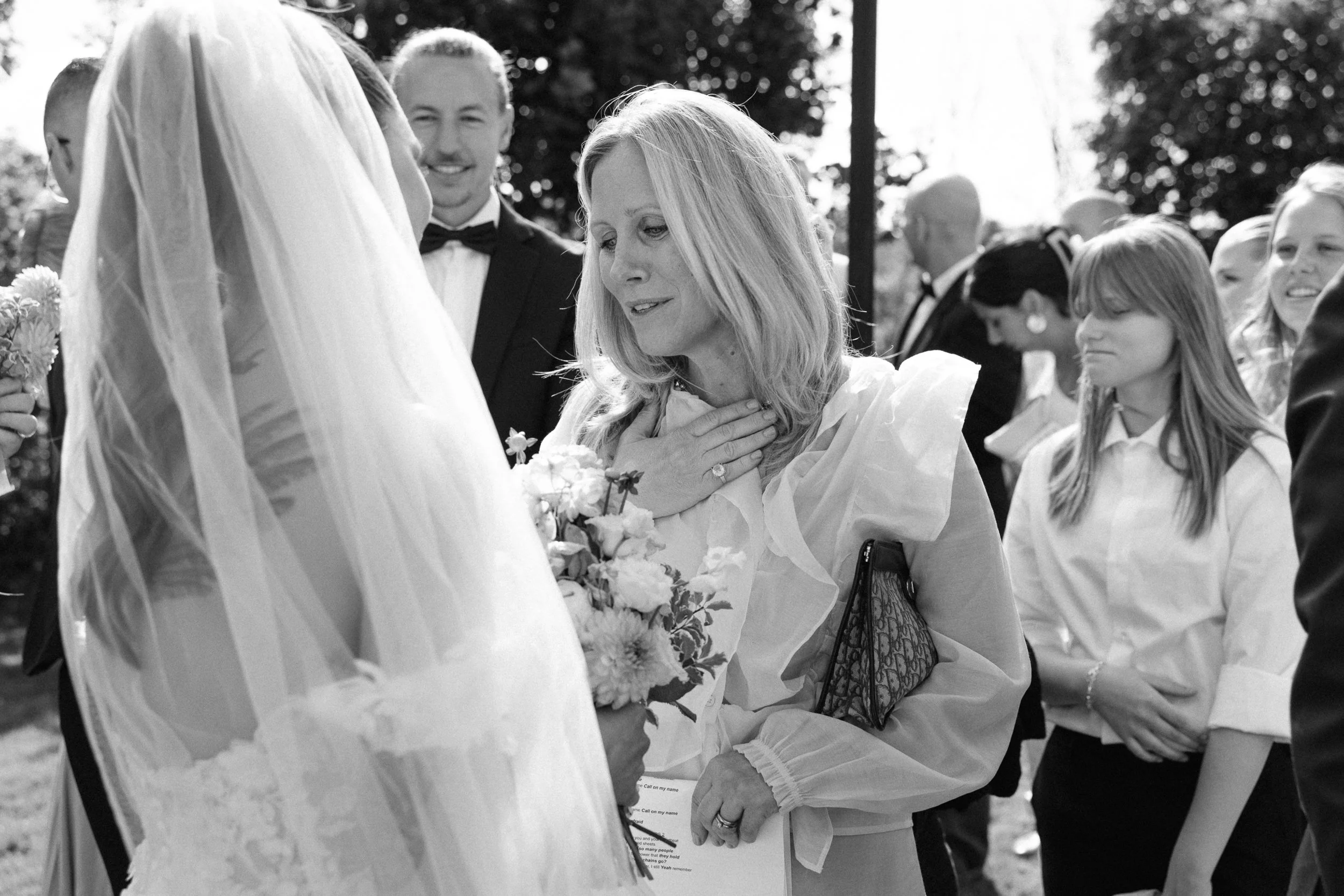 Kvinnor som deltar i ett bröllopsceremoni utomhus, med en brud i vit klänning och en kvinna som håller i blommor och bär en ljus blus, omgiven av andra gäster.  Av Sofia Wilhelmina bröllopsfotograf i Stockholm.
