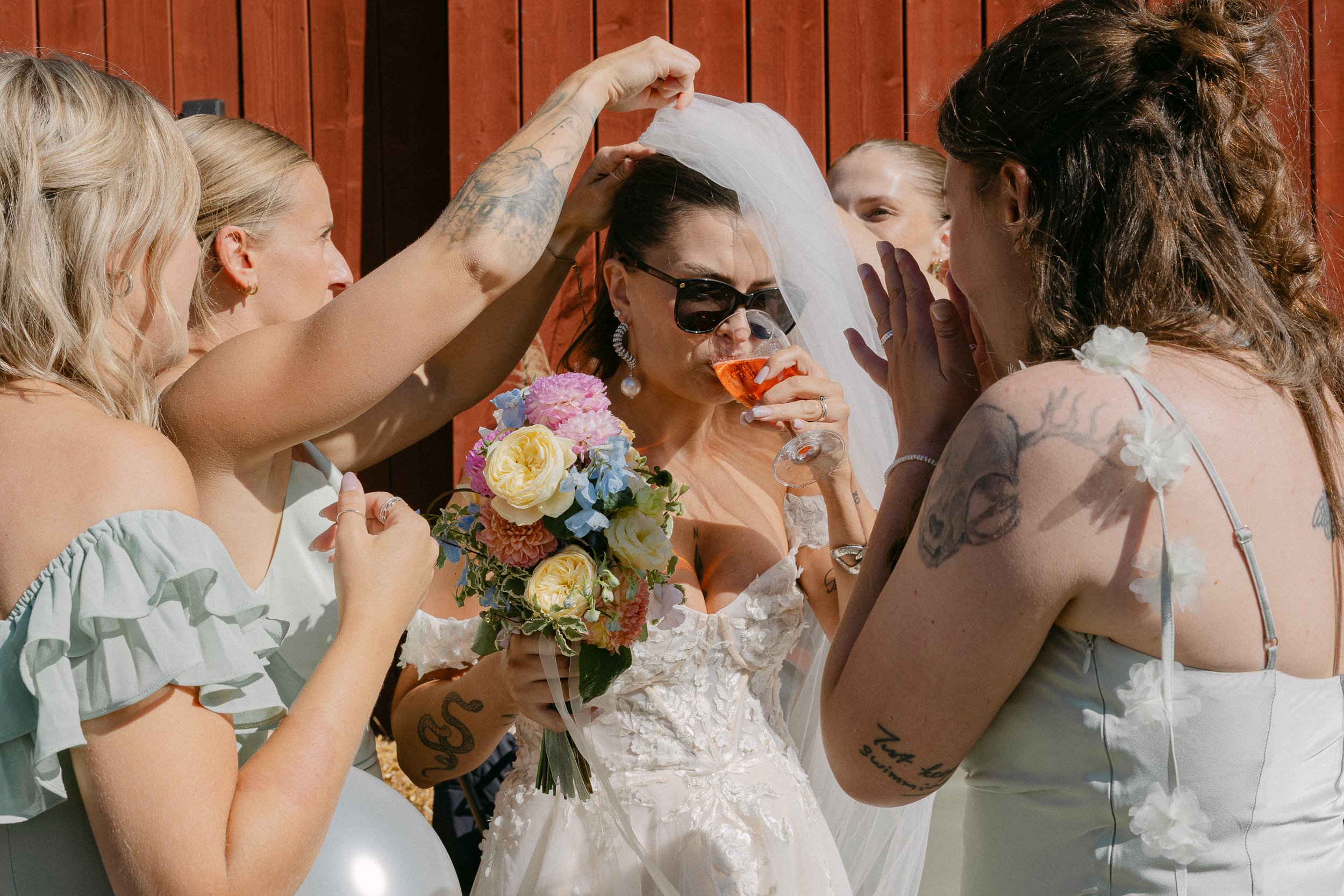 Kvinna på bröllop med solglasögon dricker vin medan andra kvinnor omkring henne hjälper till med hennes brudklänning, en av kvinnorna håller en bukett blommor.  Av Sofia Wilhelmina bröllopsfotograf i Stockholm.